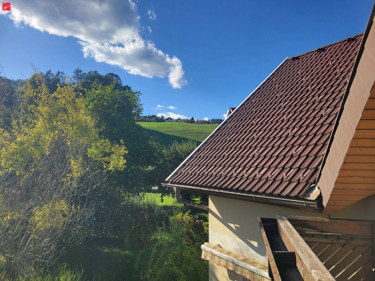 Idyllischer Ausblick auf eine grüne Hügellandschaft und das Ziegeldach eines Hauses.