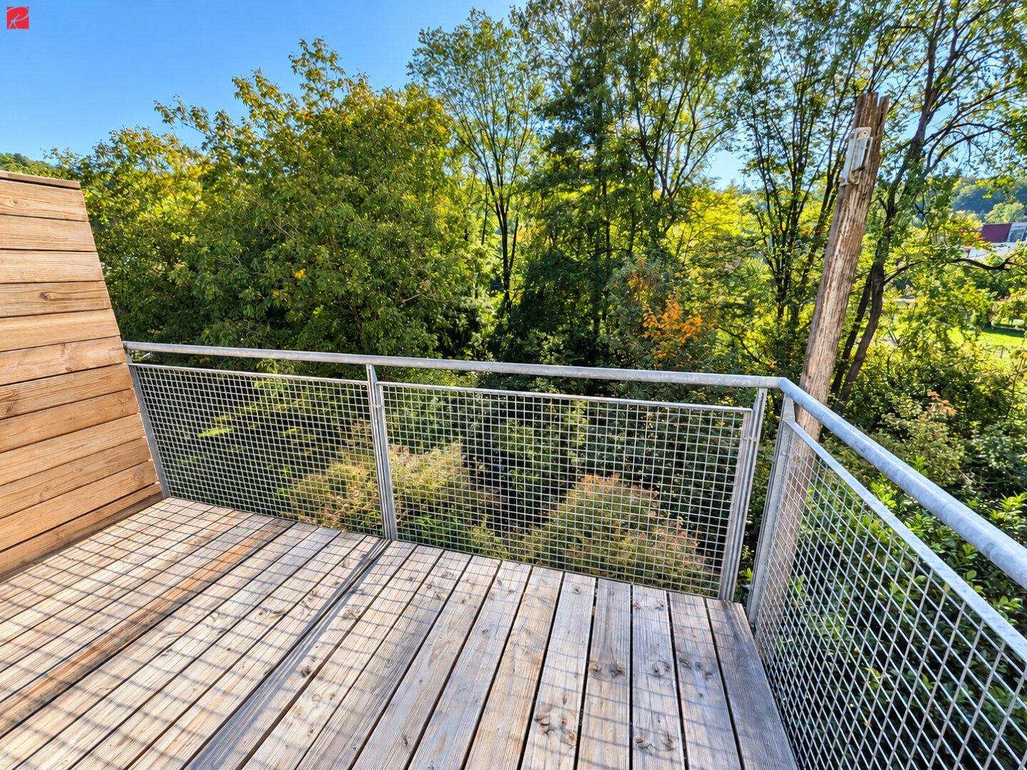 Heller Holzbalkon mit Metallgeländer und weitem Blick auf grüne Bäume und die Umgebung.