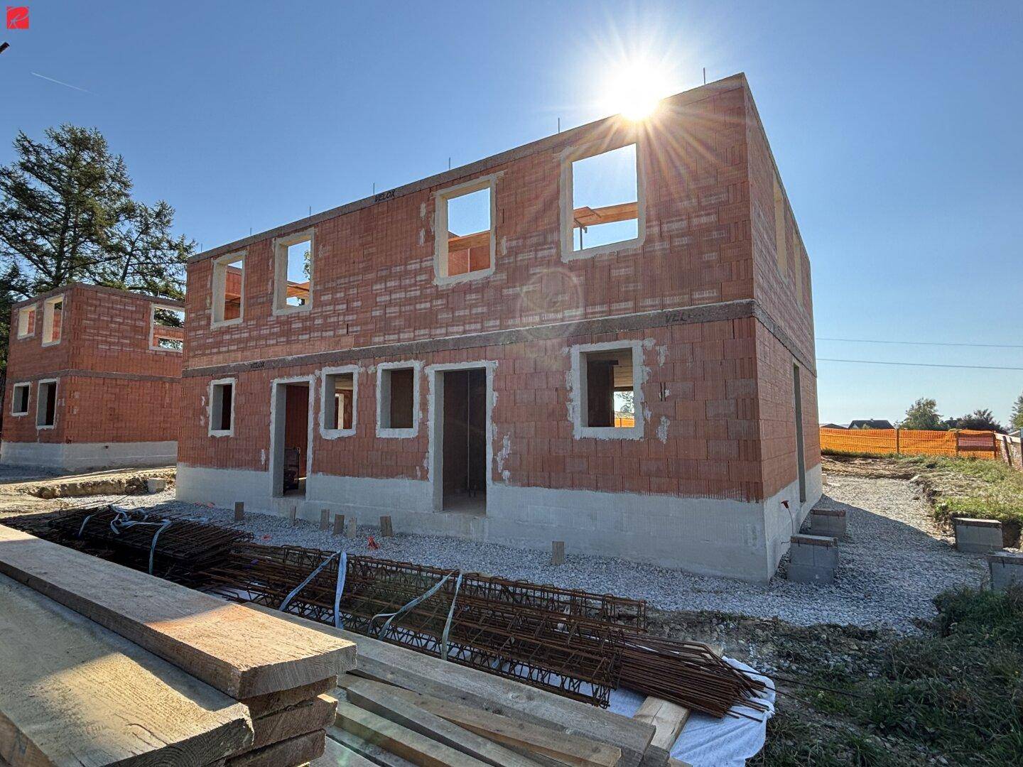 Ein im Bau befindliches Ziegelhaus mit Rohbau und Baumaterialien unter strahlendem Sonnenschein.