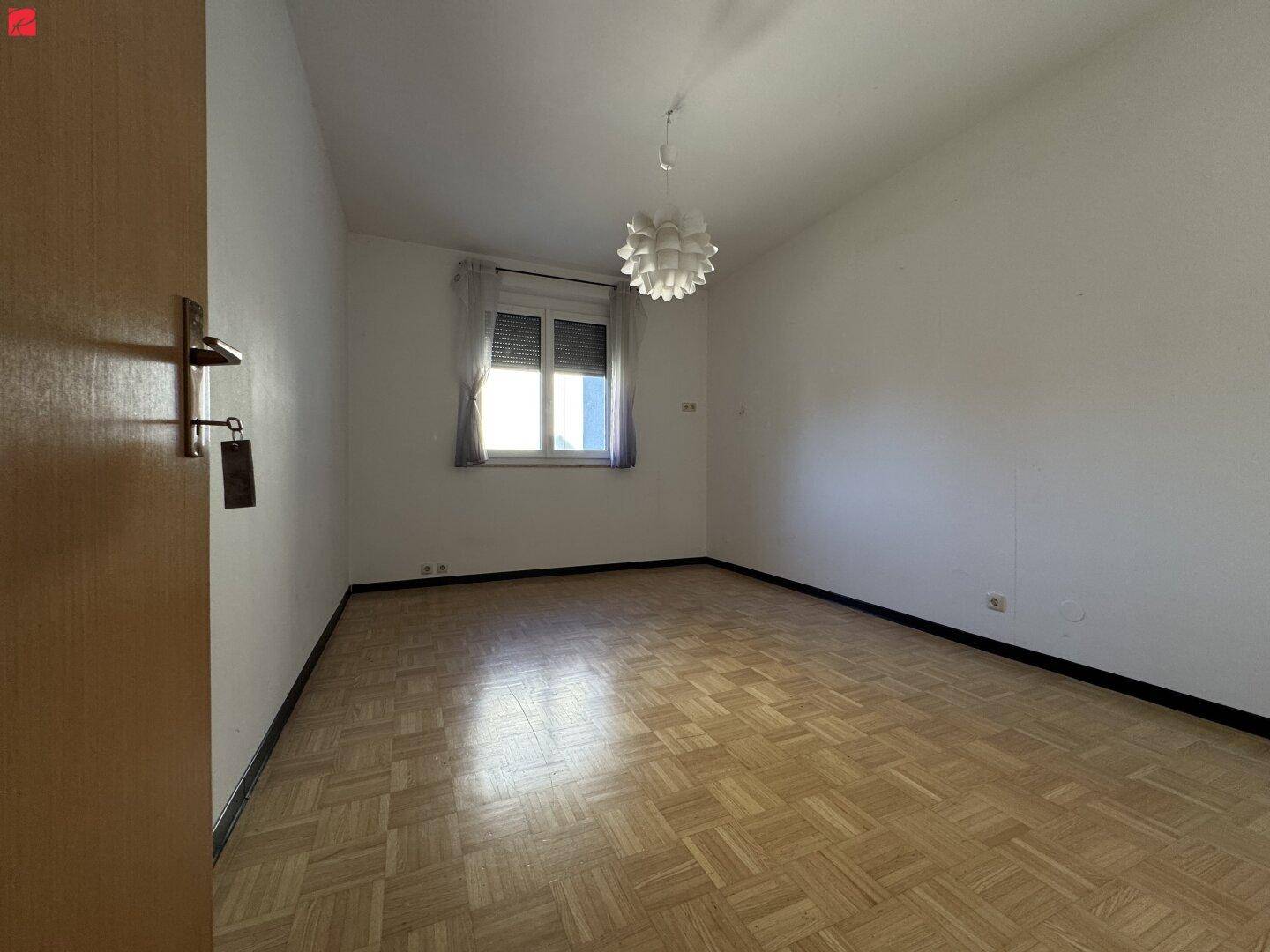 Schlafzimmer mit Parkettboden, Fenster mit Rollladen und einer modernen Deckenleuchte, die den Raum erhellt.