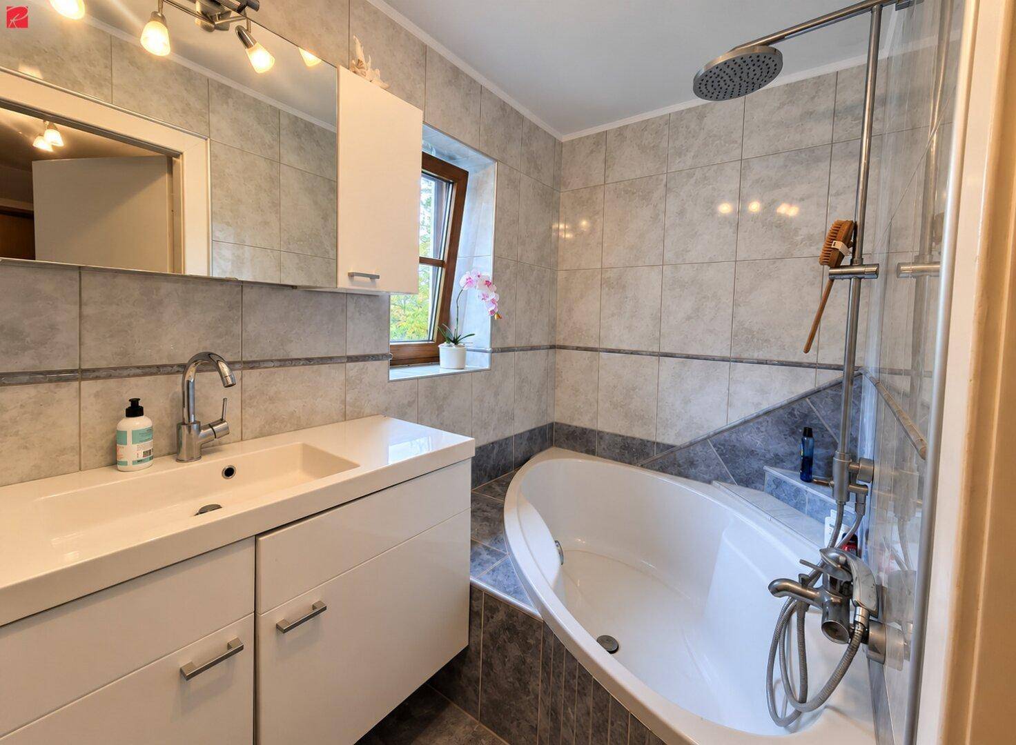 Gefliestes Badezimmer mit Badewanne, Regendusche, Einzelwaschbecken und Fenster für Tageslicht.