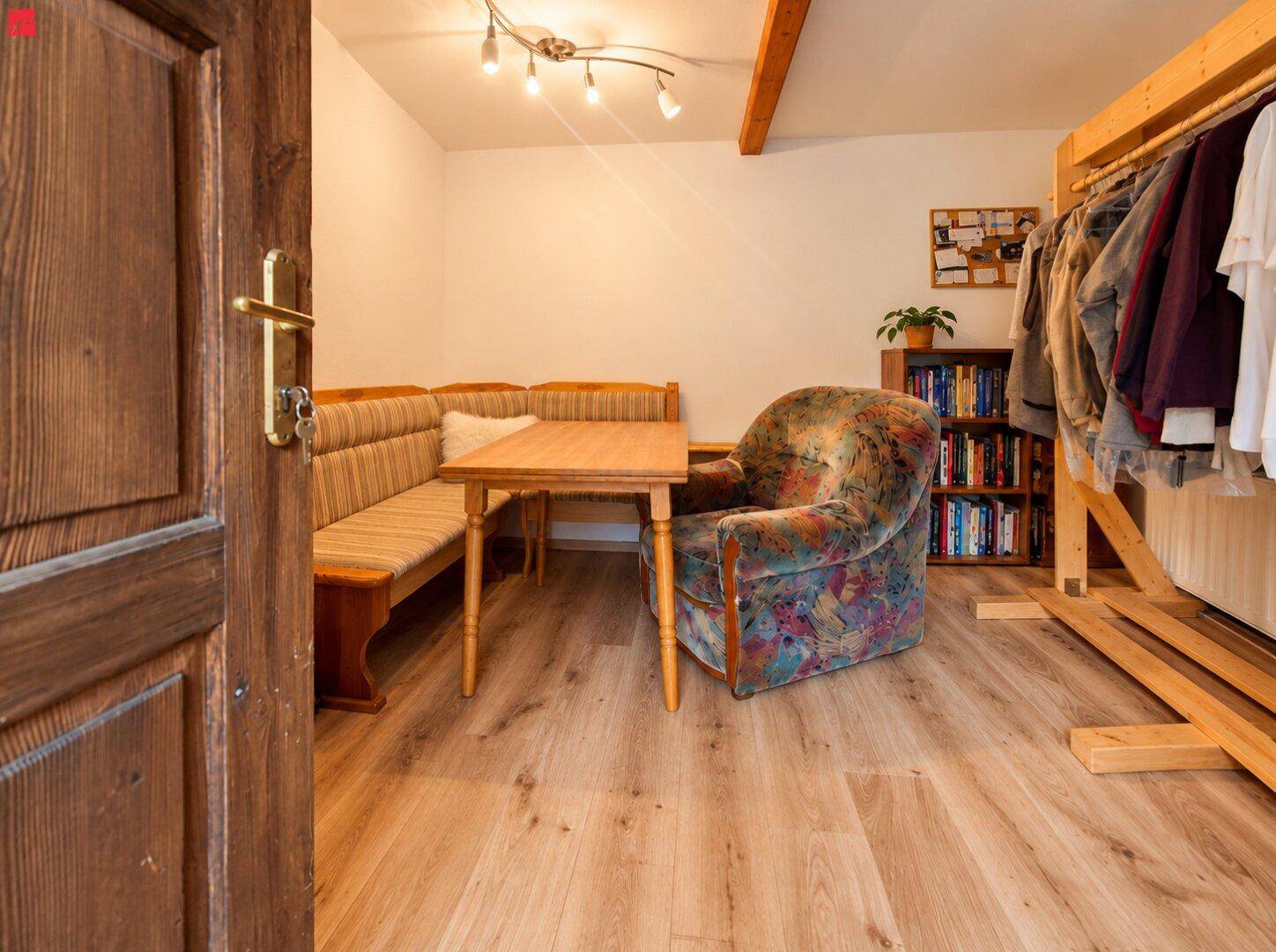Essbereich mit Holztisch, Eckbank, Sessel und sichtbaren Deckenbalken, daneben ein Kleiderständer.