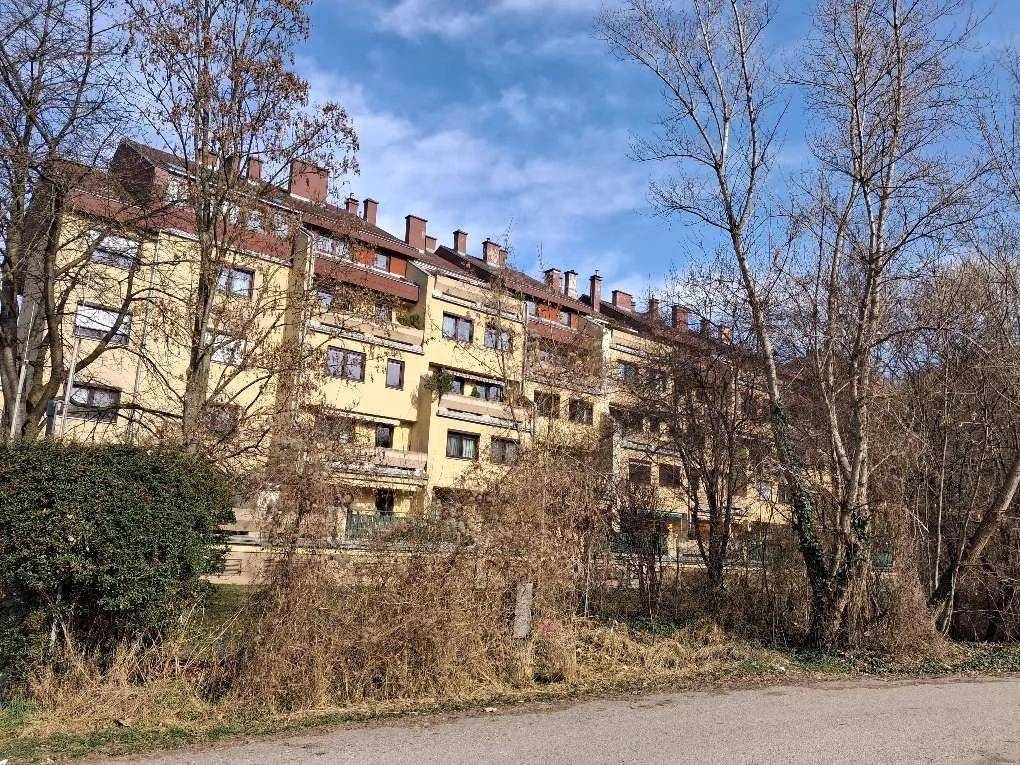 Eine lange Wohnanlage mit mehreren Balkonen, umgeben von Bäumen und Sträuchern unter blauem Himmel.