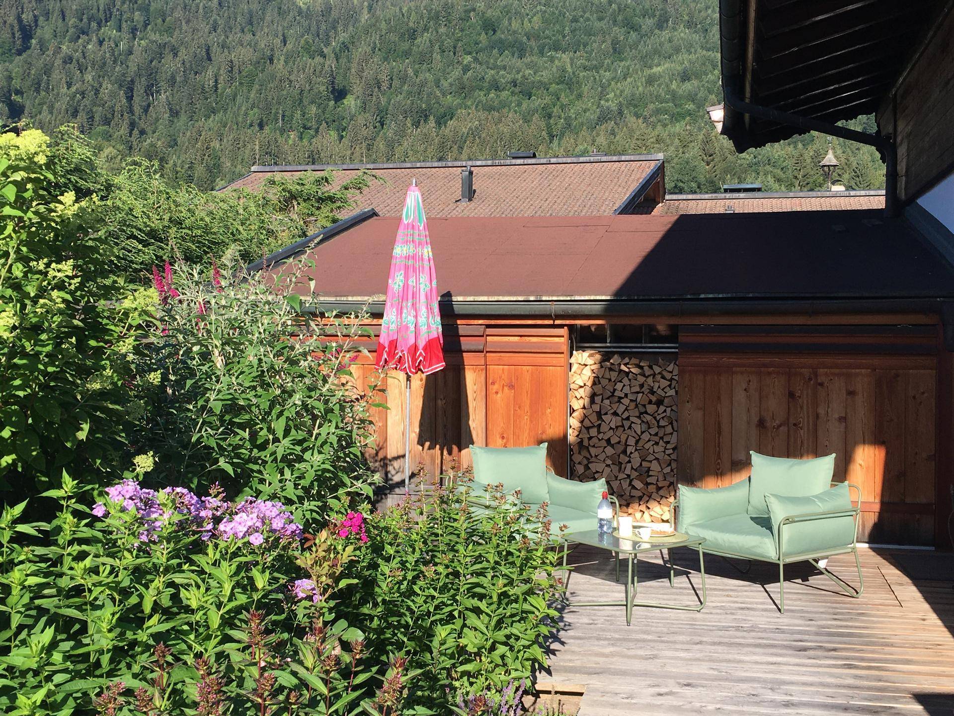 Gemütlicher Sitzbereich auf einer Holzterrasse, umgeben von Blumen und mit Blick auf die bewaldeten Berge.