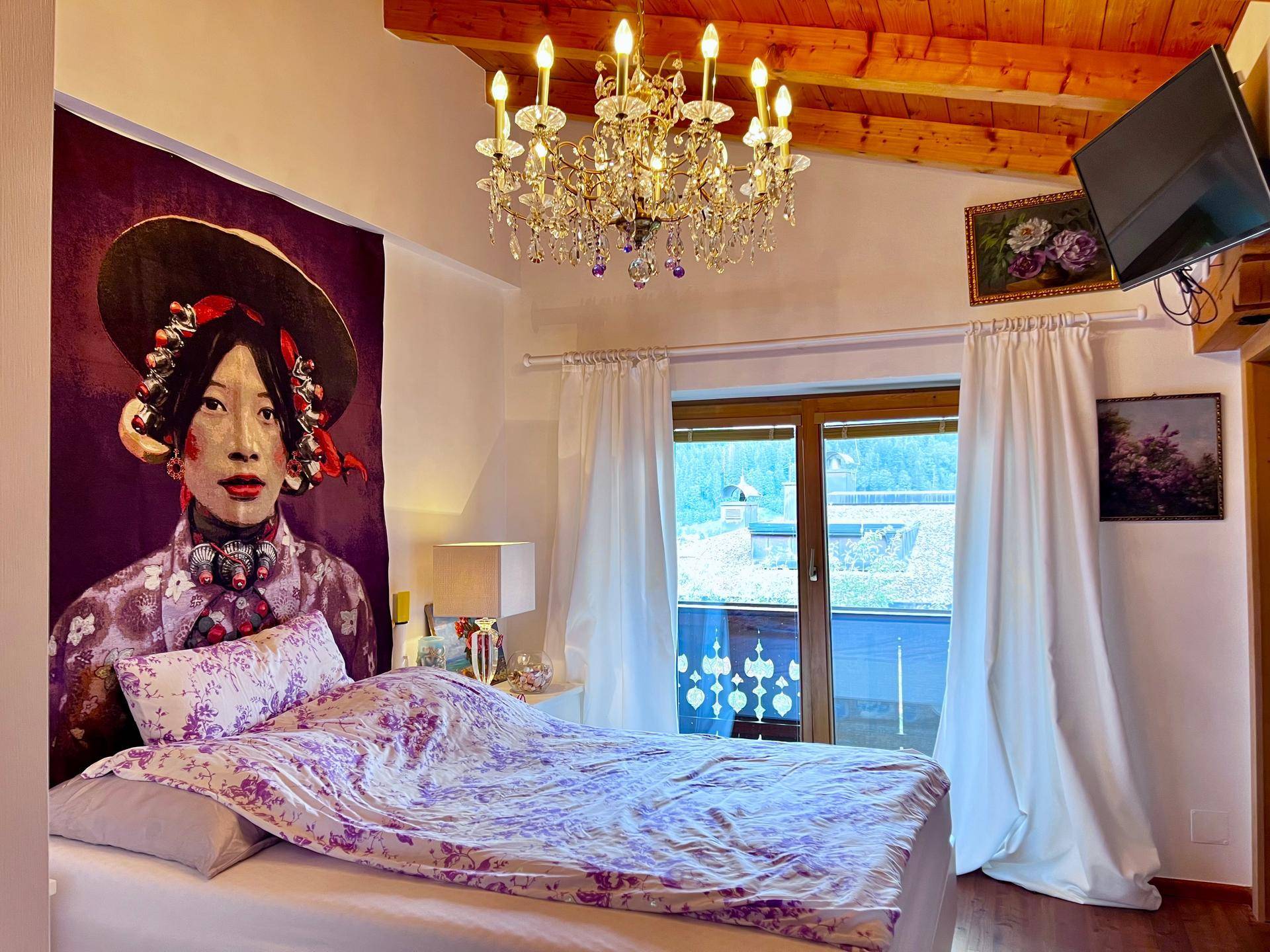 Gemütliches Schlafzimmer mit Doppelbett, Kronleuchter, Holzbalkendecke und Zugang zum Balkon mit Aussicht.