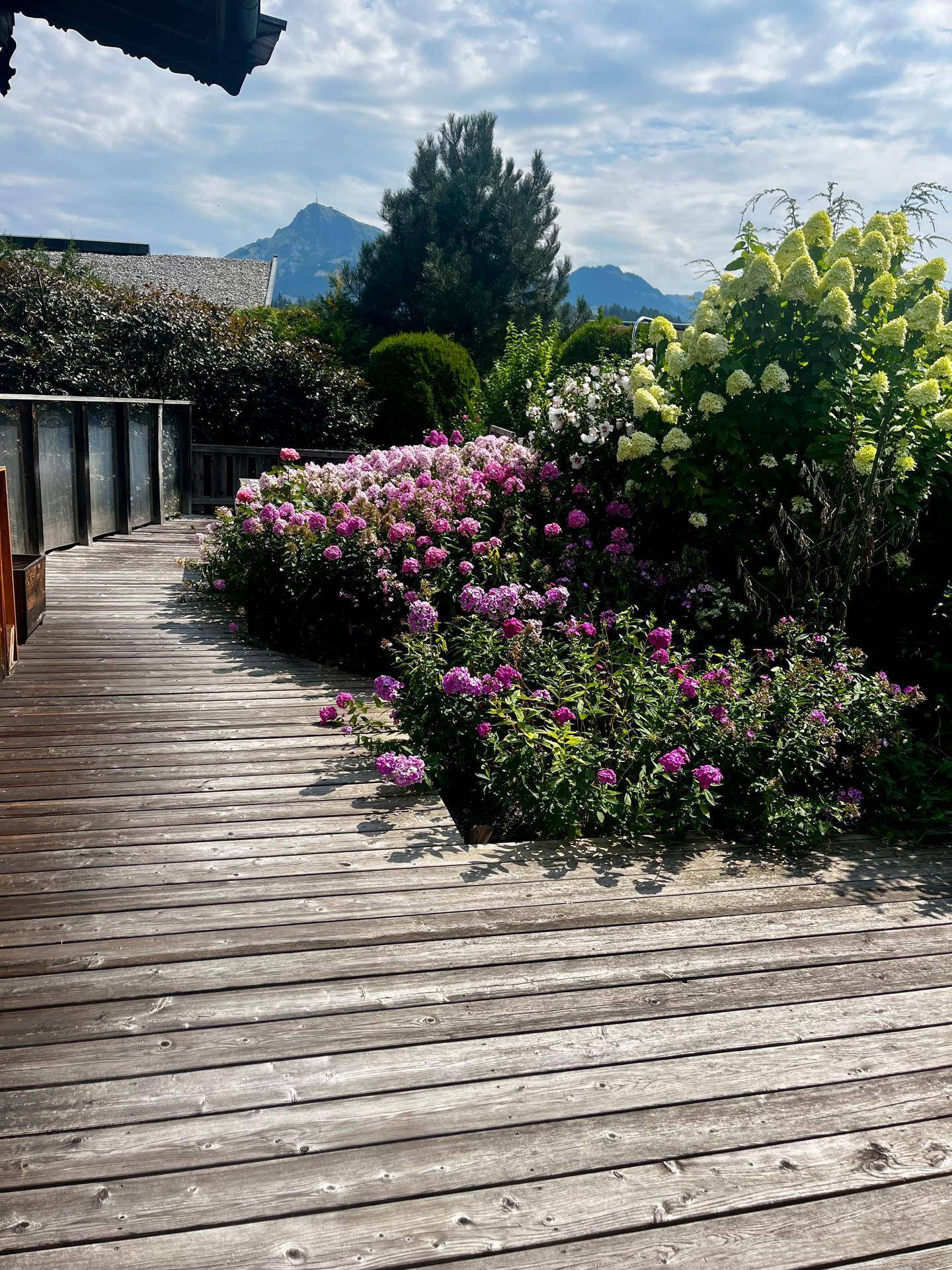 Geräumige Holzterrasse umgeben von bunten Blumen und mit Blick auf die majestätischen Berge.