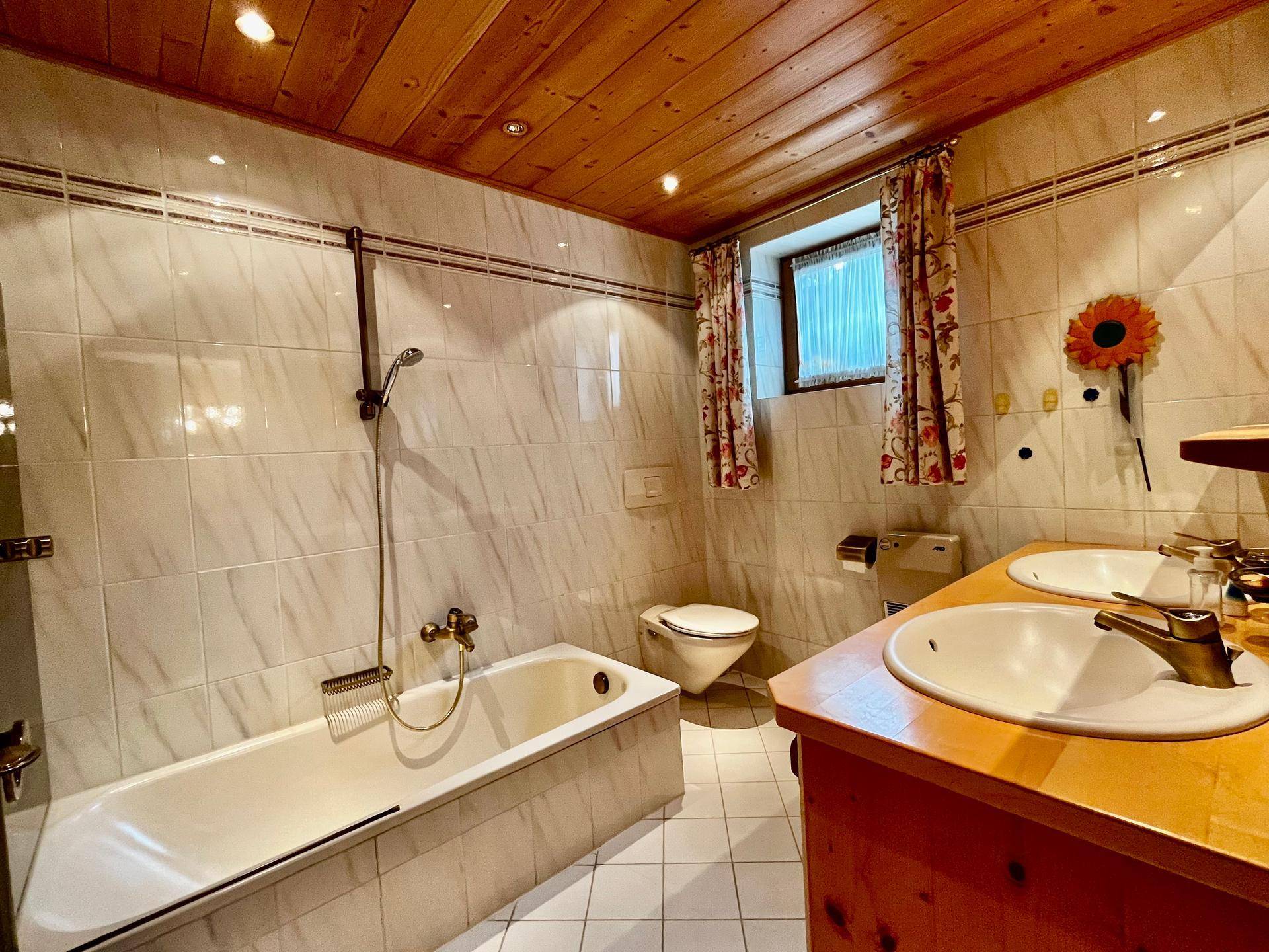 Helles Badezimmer mit weißen Fliesen, Badewanne, Doppelwaschbecken und einem kleinen Fenster.