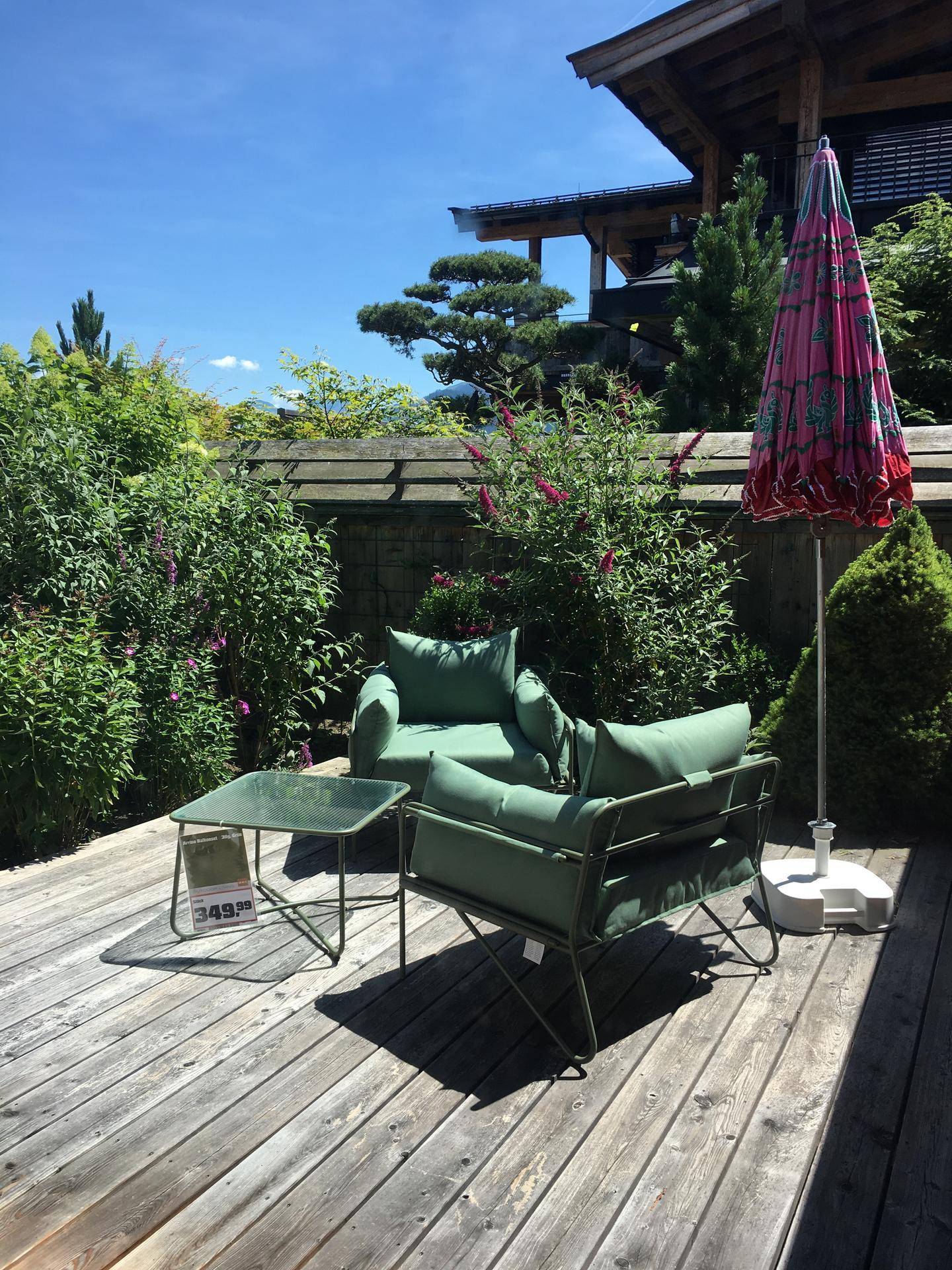 Sonnige Holzterrasse mit bequemen grünen Gartenmöbeln, umgeben von üppiger Bepflanzung.