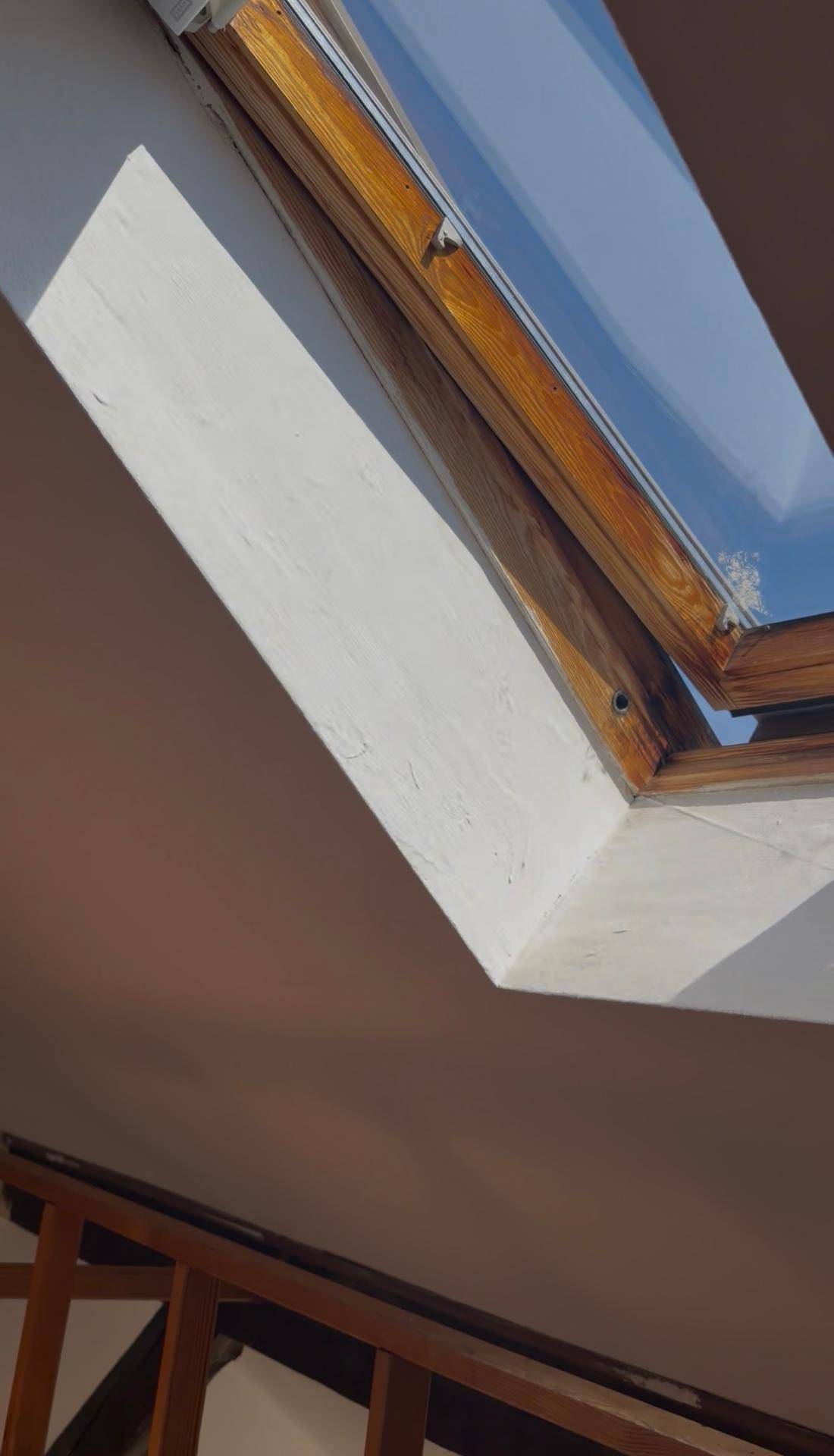 Nahaufnahme eines Holz-Dachfensters in einer weißen Dachschräge, das den blauen Himmel zeigt.