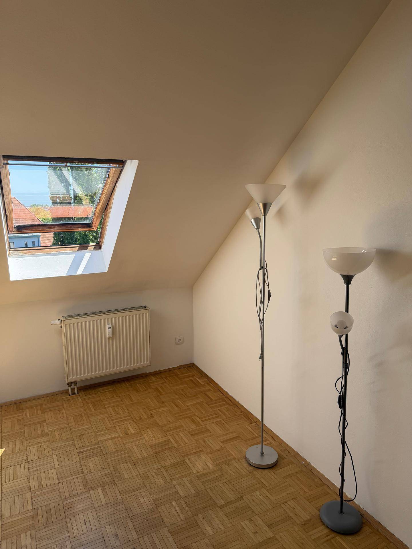 Lichtdurchfluteter Raum mit Dachschräge, Dachfenster, Heizkörper und zwei Stehlampen.