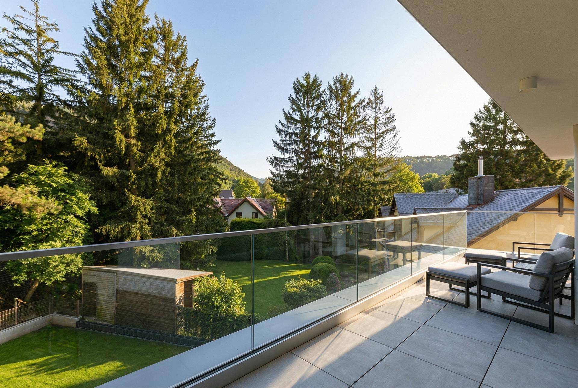 Sonniger Balkon mit Glasbrüstung, Sitzgelegenheiten und weitem Blick ins Grüne.