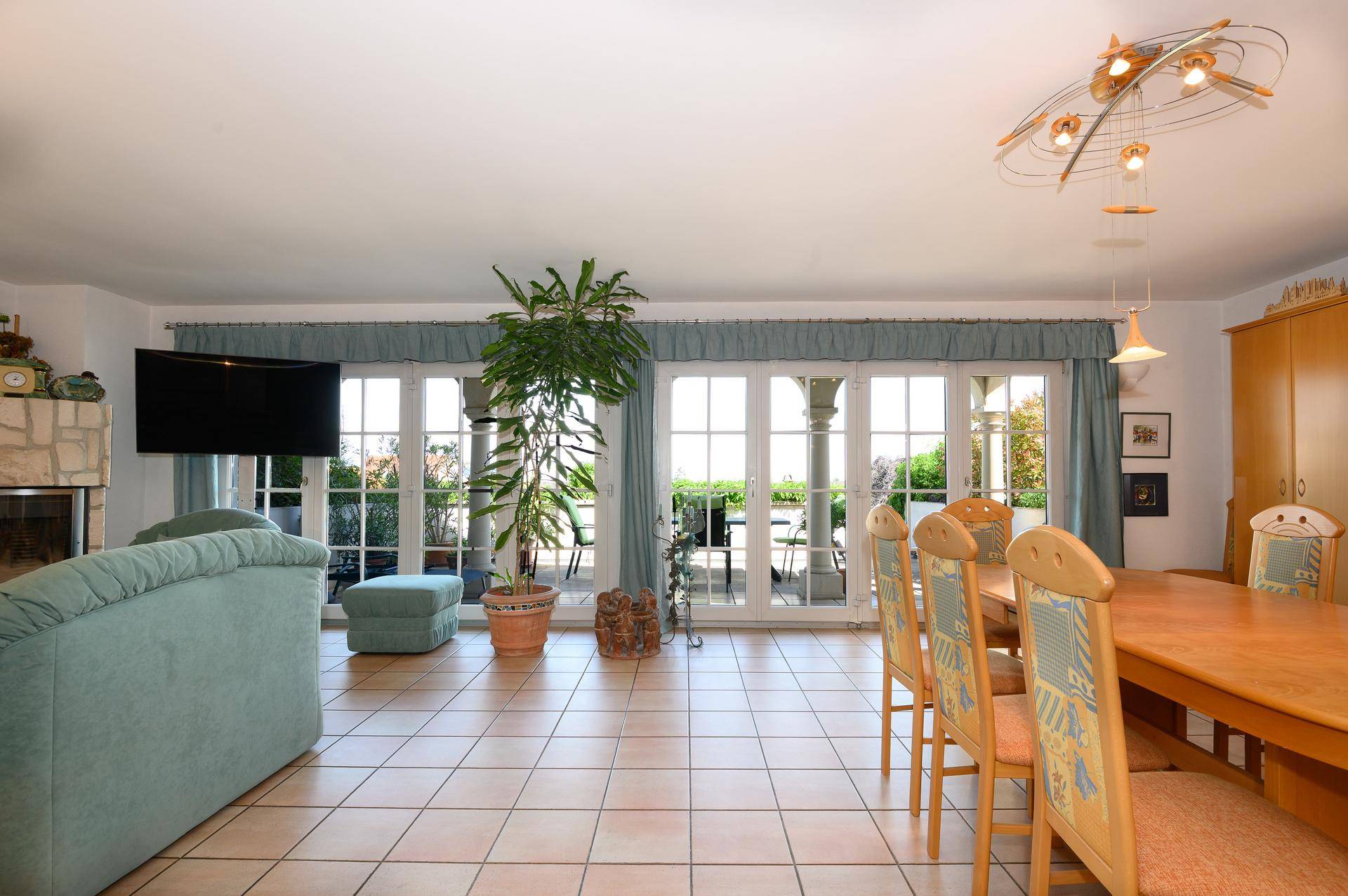 Geräumiger Wohn-Essbereich mit Fliesenboden, Kamin und großen Fenstertüren, die auf eine Terrasse führen.
