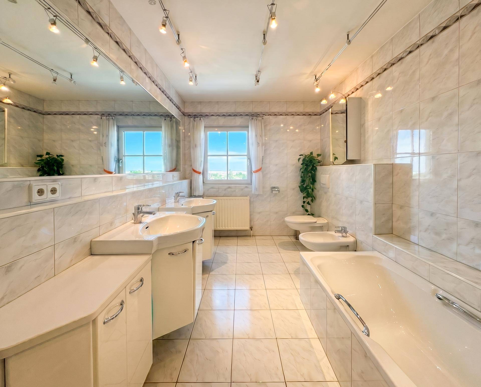 Helles Badezimmer mit weißen Fliesen, Doppelwaschbecken, Badewanne, Bidet und Fenster für Tageslicht.
