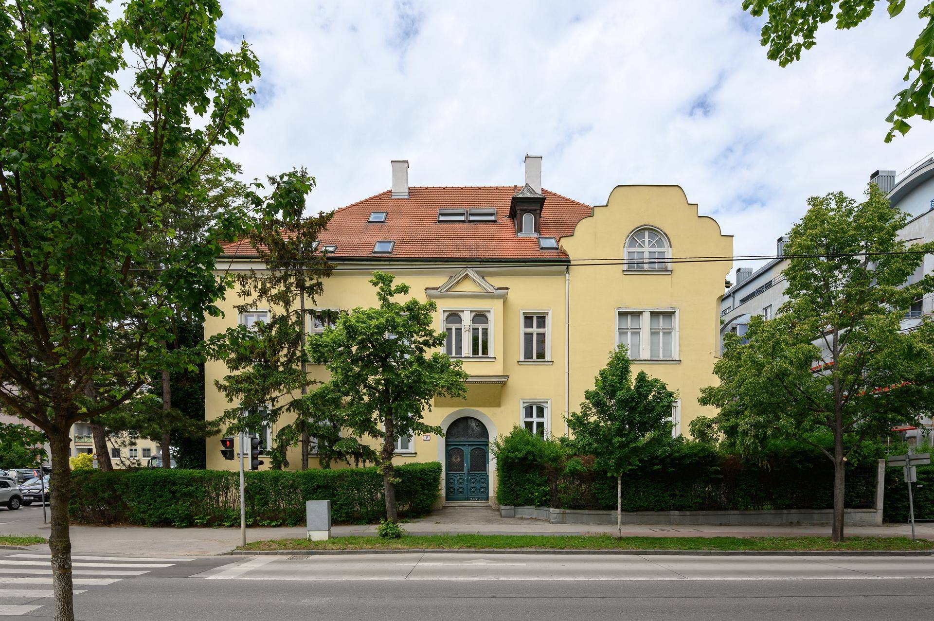 Gelbes Gebäude mit rotem Ziegeldach und grüner Bepflanzung an einer Straße.