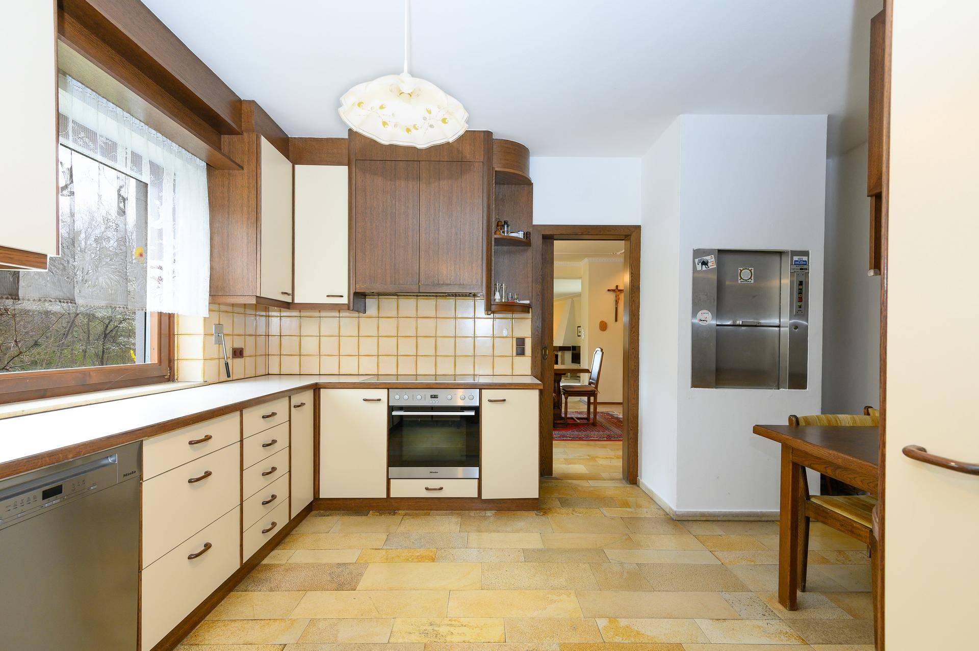 Funktionale Küche mit Holzschränken, Fliesenboden, integriertem Geschirrspüler und Speiseaufzug.