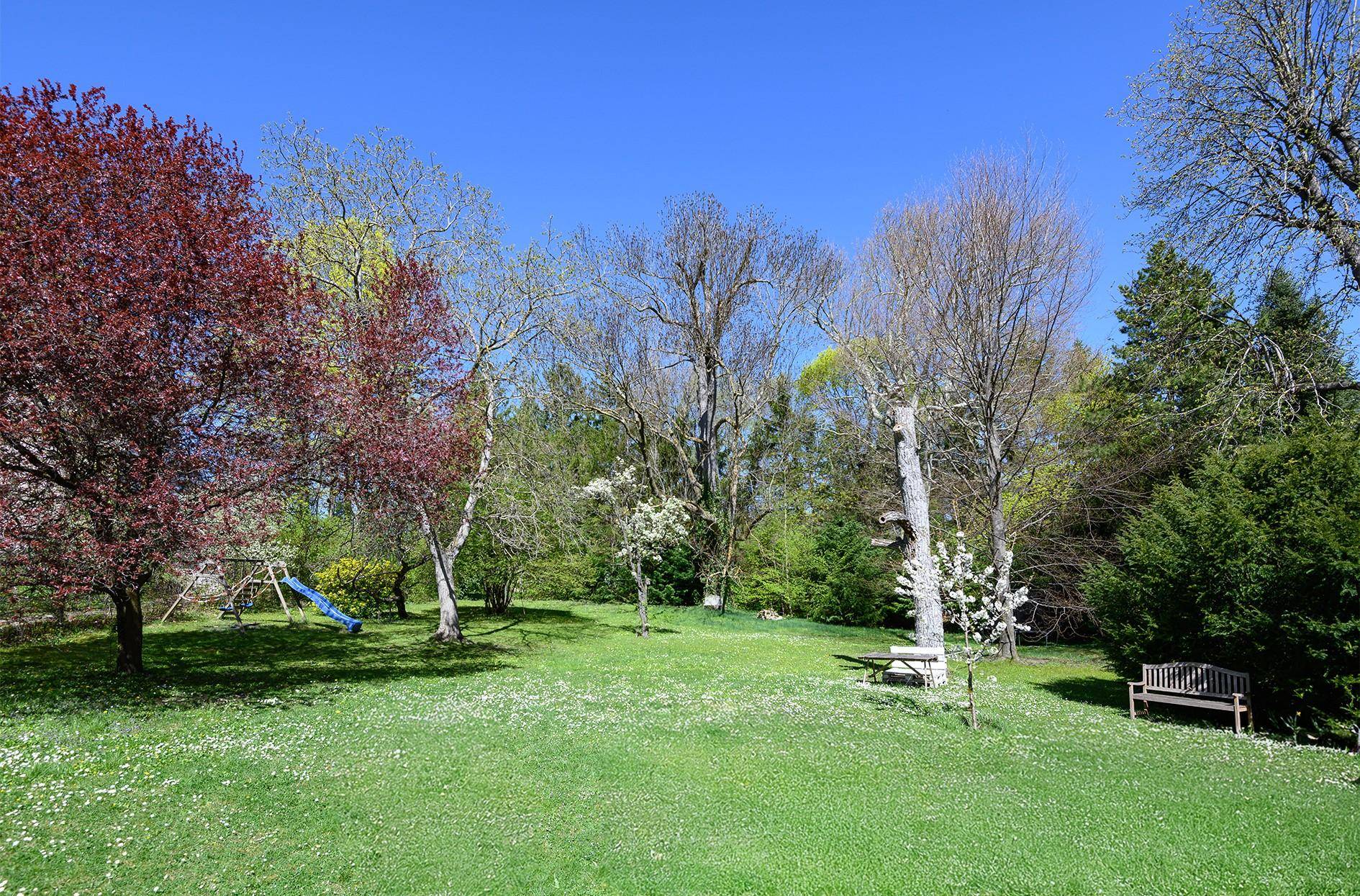 Großer, sonniger Garten mit Rasenfläche, alten Bäumen und einem Kinderspielgerät.