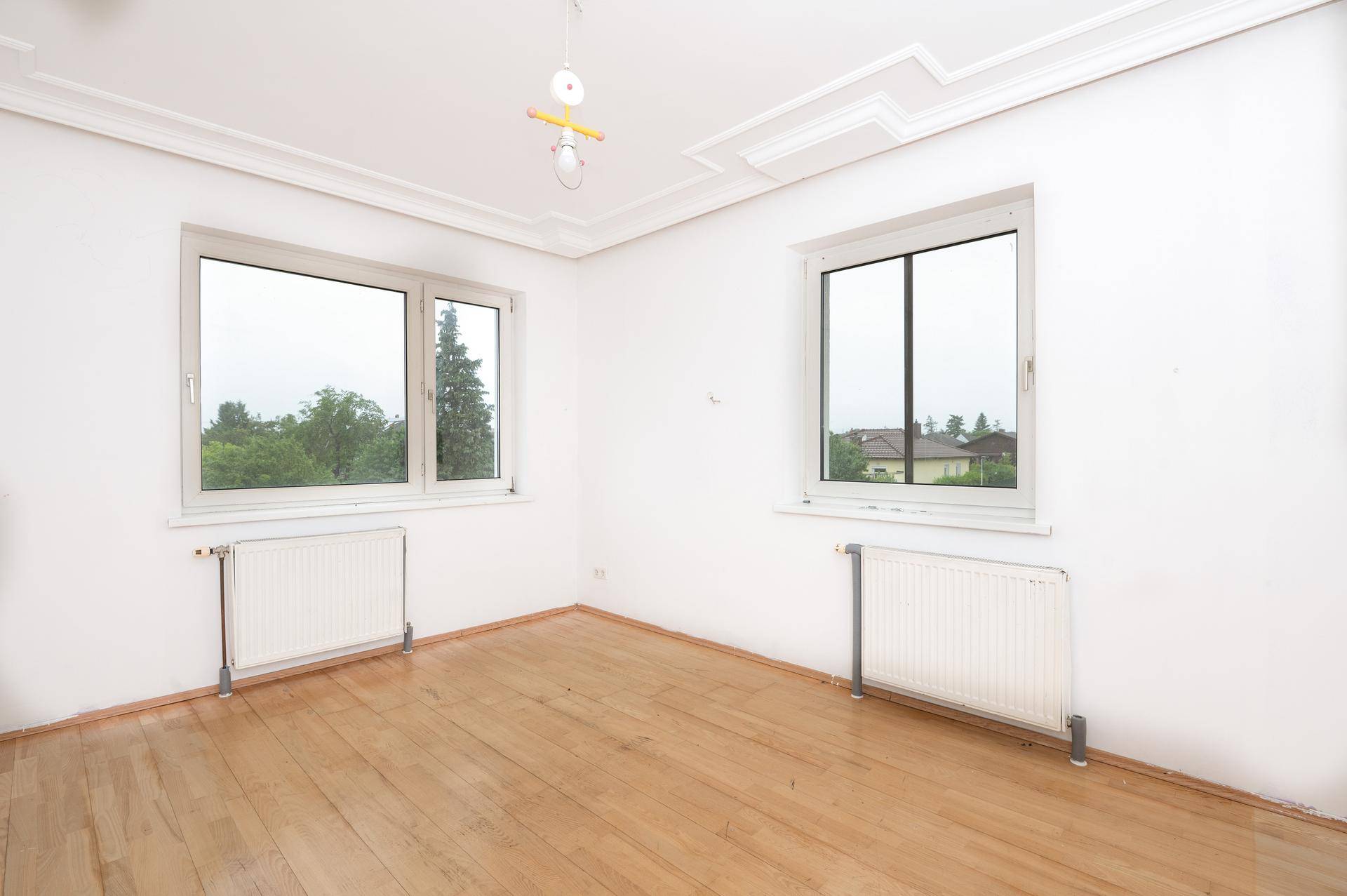 Helles Zimmer mit Holzdielenboden, zwei Fenstern und dekorativen Stuckelementen an der Decke.