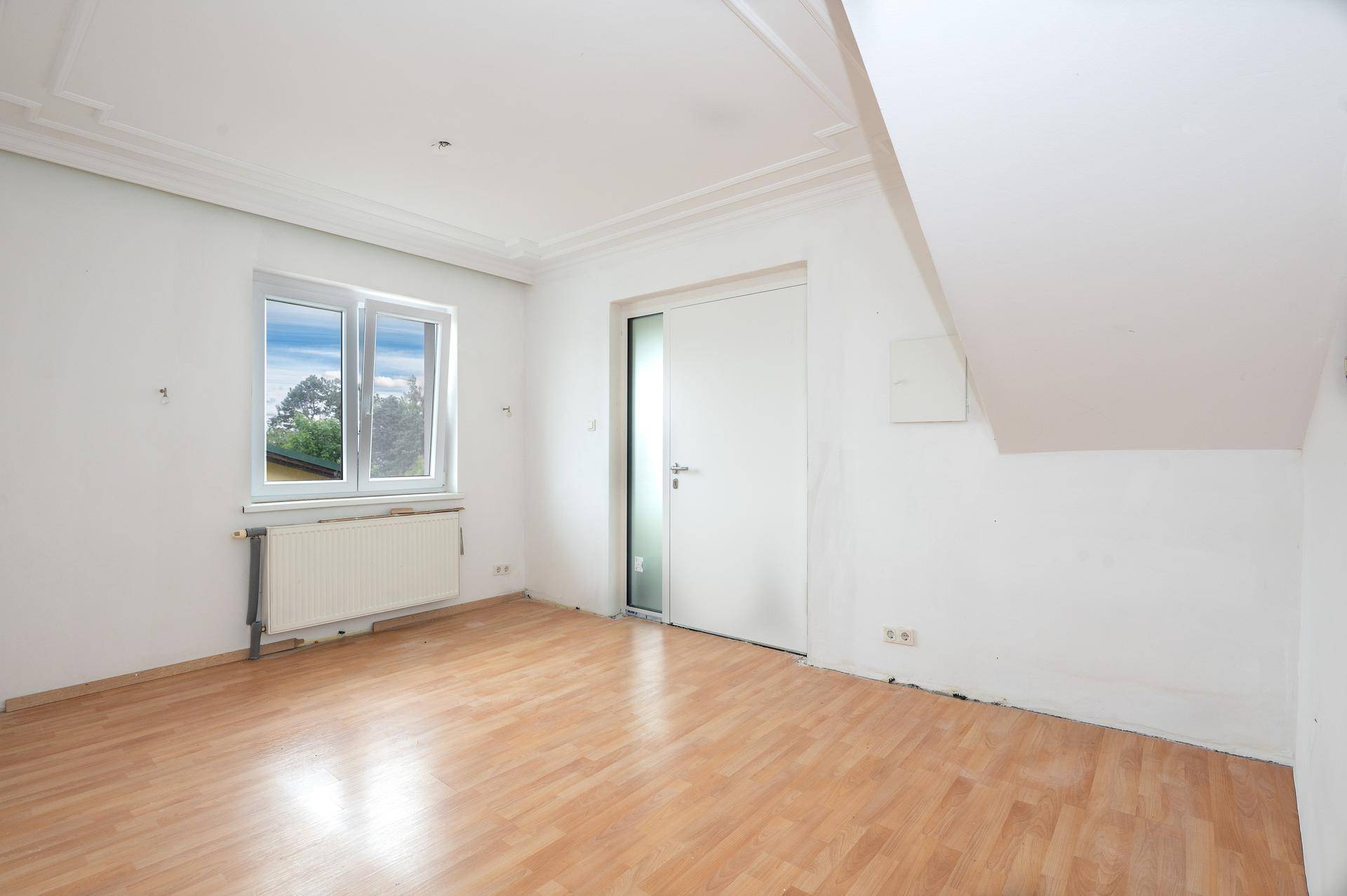 Leeres Zimmer mit hellem Laminatboden, weiß gestrichenen Wänden und einem Fenster.