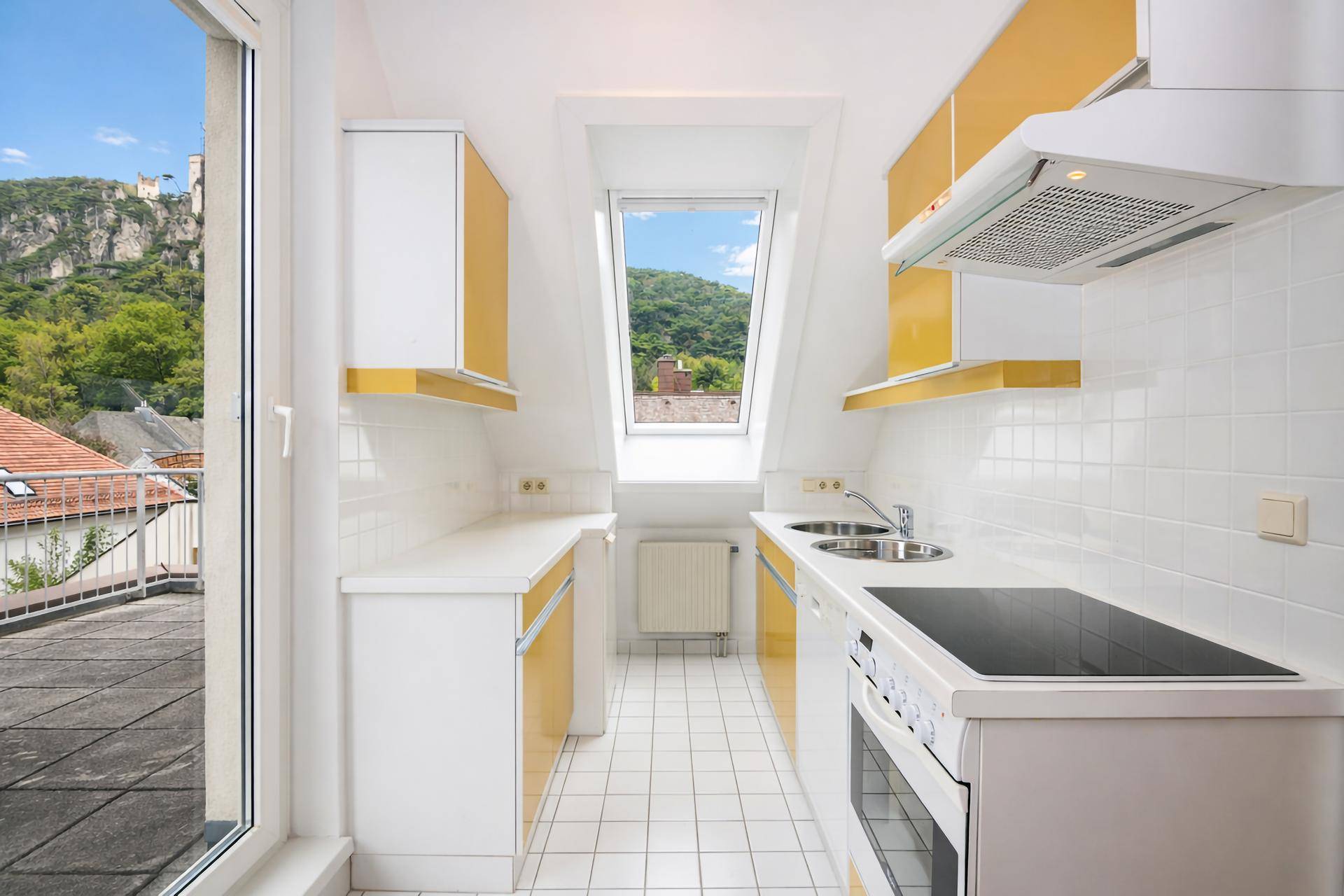 Funktionale Küche mit weißen und gelben Schränken, Fliesenboden und Zugang zur Terrasse mit Burgblick.