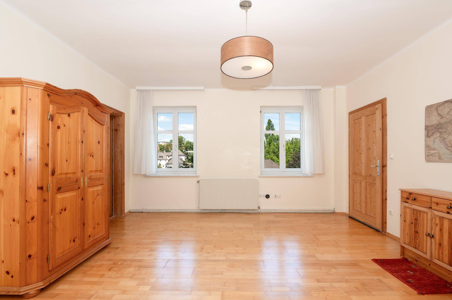 Leeres Zimmer mit Holzboden, zwei Fenstern und einem großen Holzschrank.
