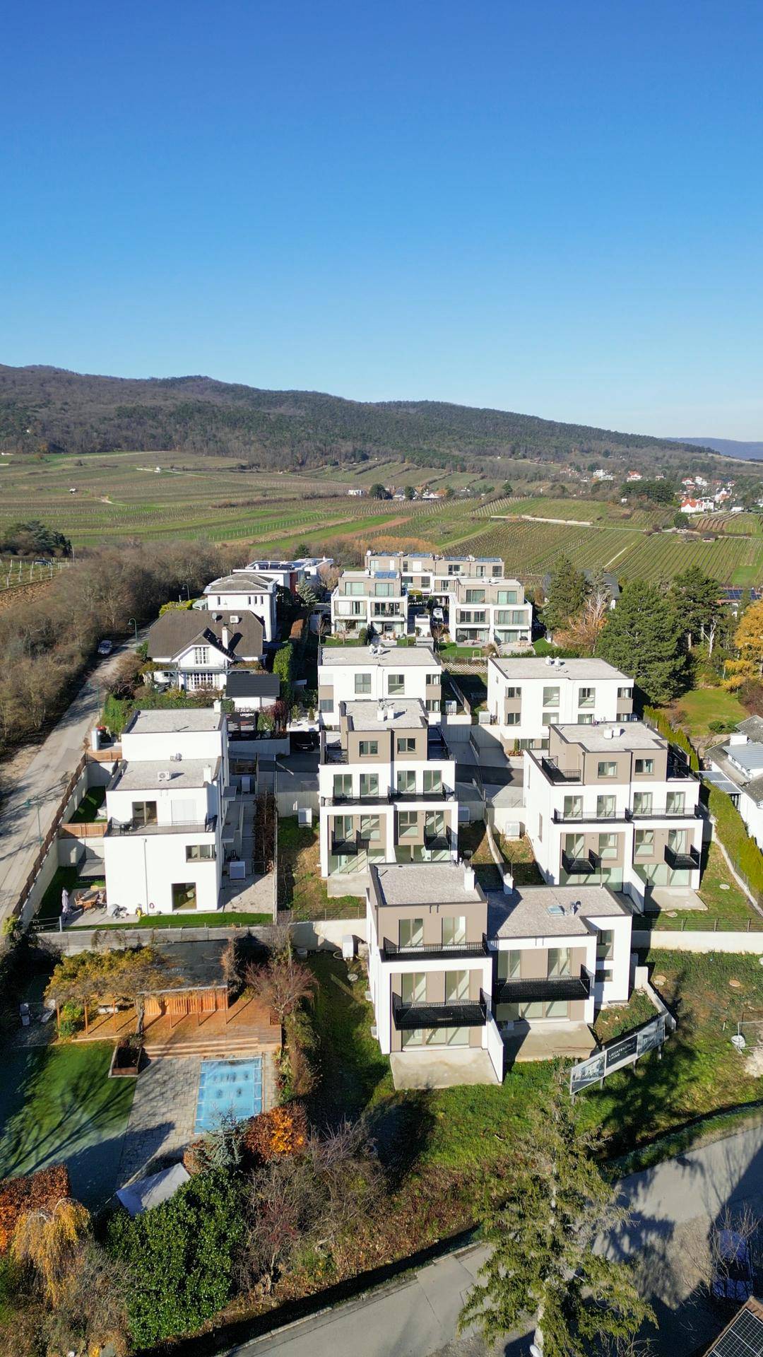 Luftaufnahme einer modernen Wohnanlage in grüner Umgebung mit Weinbergen und Bergen im Hintergrund.