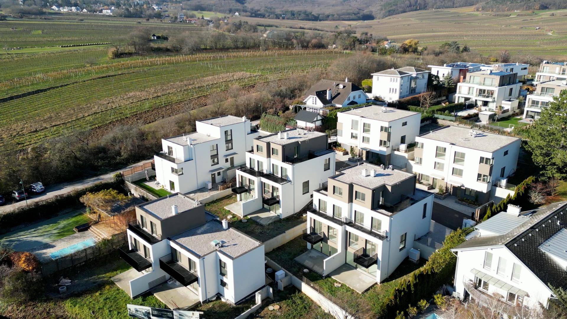 Luftaufnahme einer modernen Wohnsiedlung mit weißen Gebäuden inmitten von Weinbergen und grüner Landschaft.