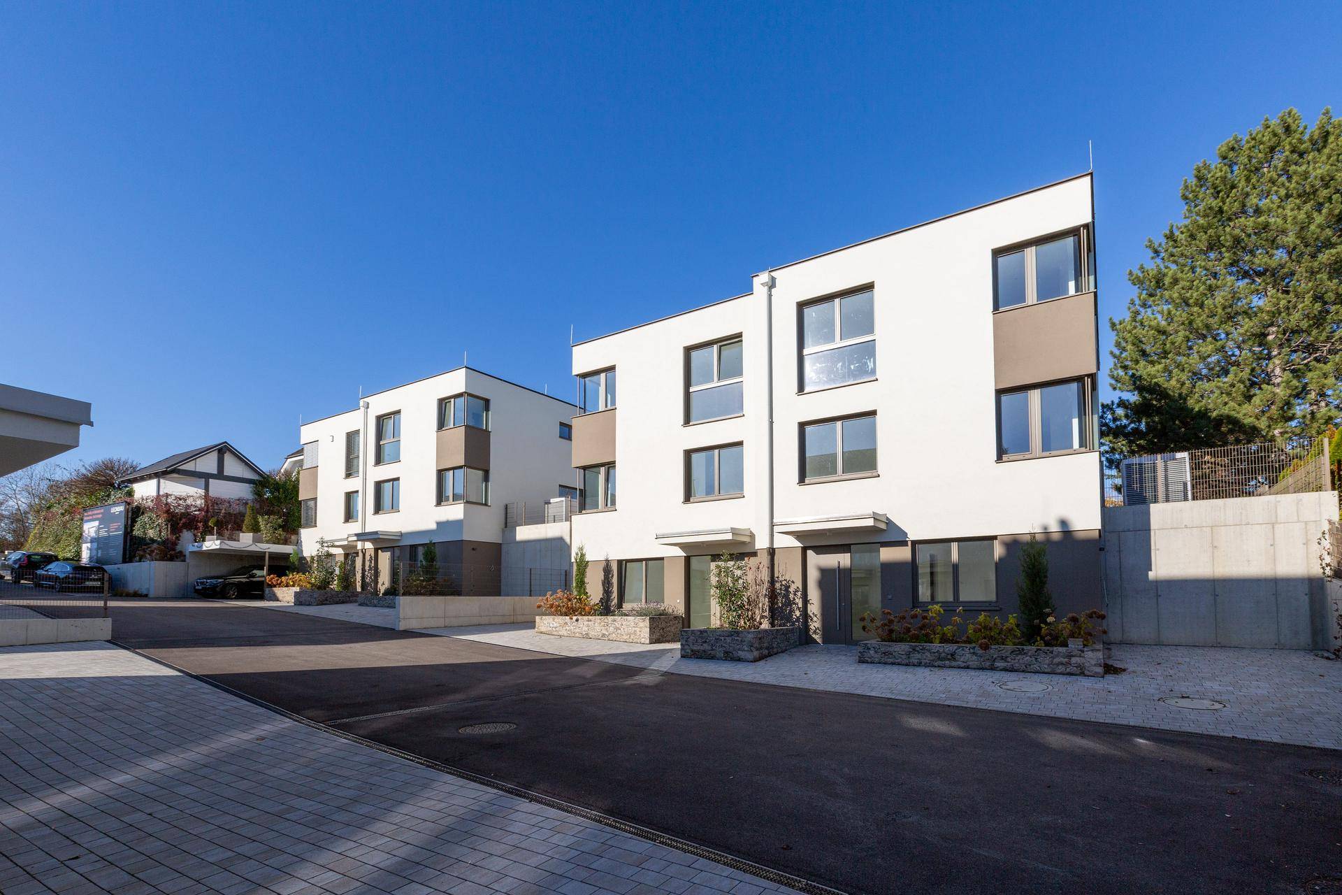 Moderne Reihenhäuser mit weißer Fassade und großen Fenstern an einer ruhigen Straße unter blauem Himmel.