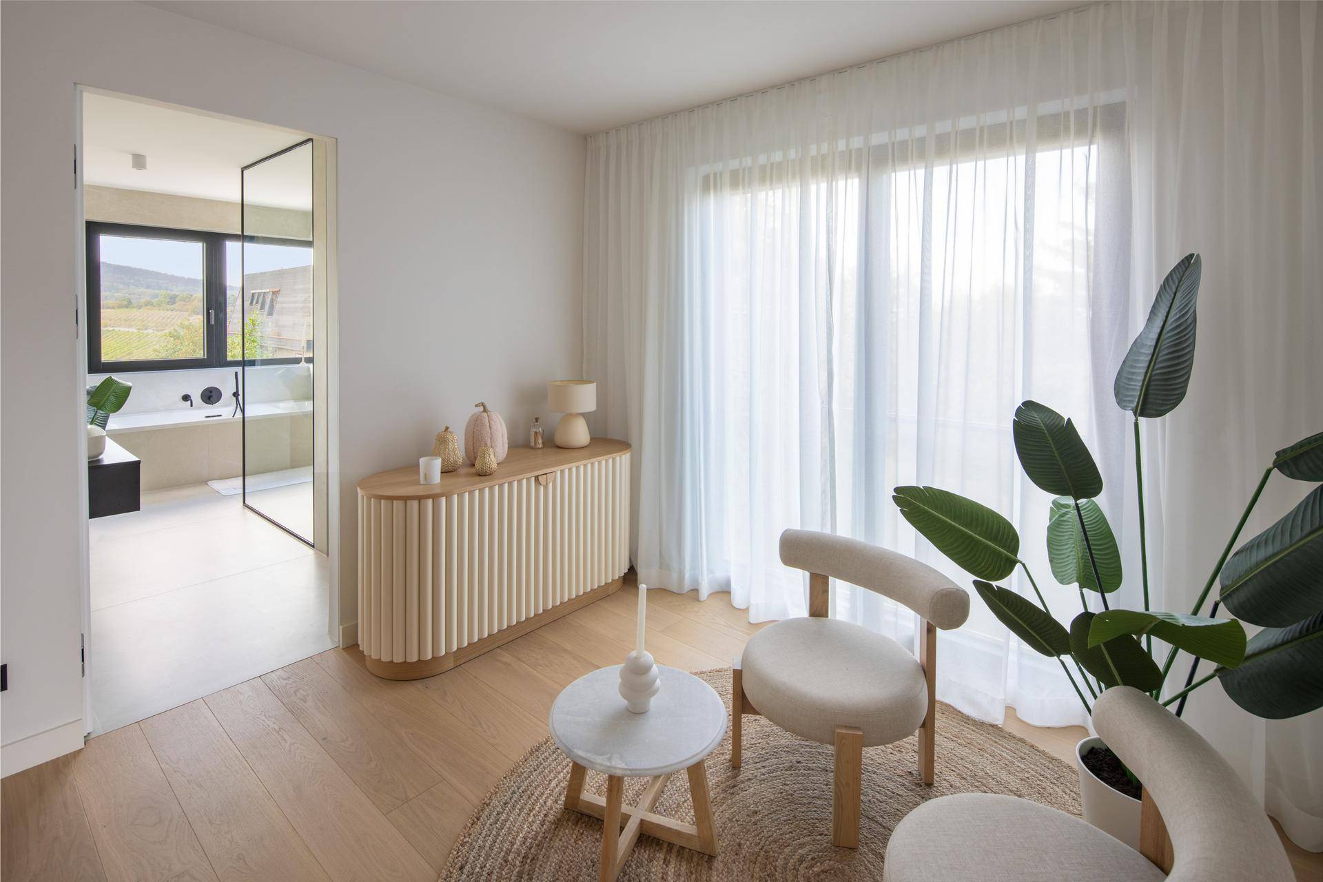 Gemütlicher Sitzbereich mit Blick in ein modernes Badezimmer und großen Fenstern mit Vorhängen.