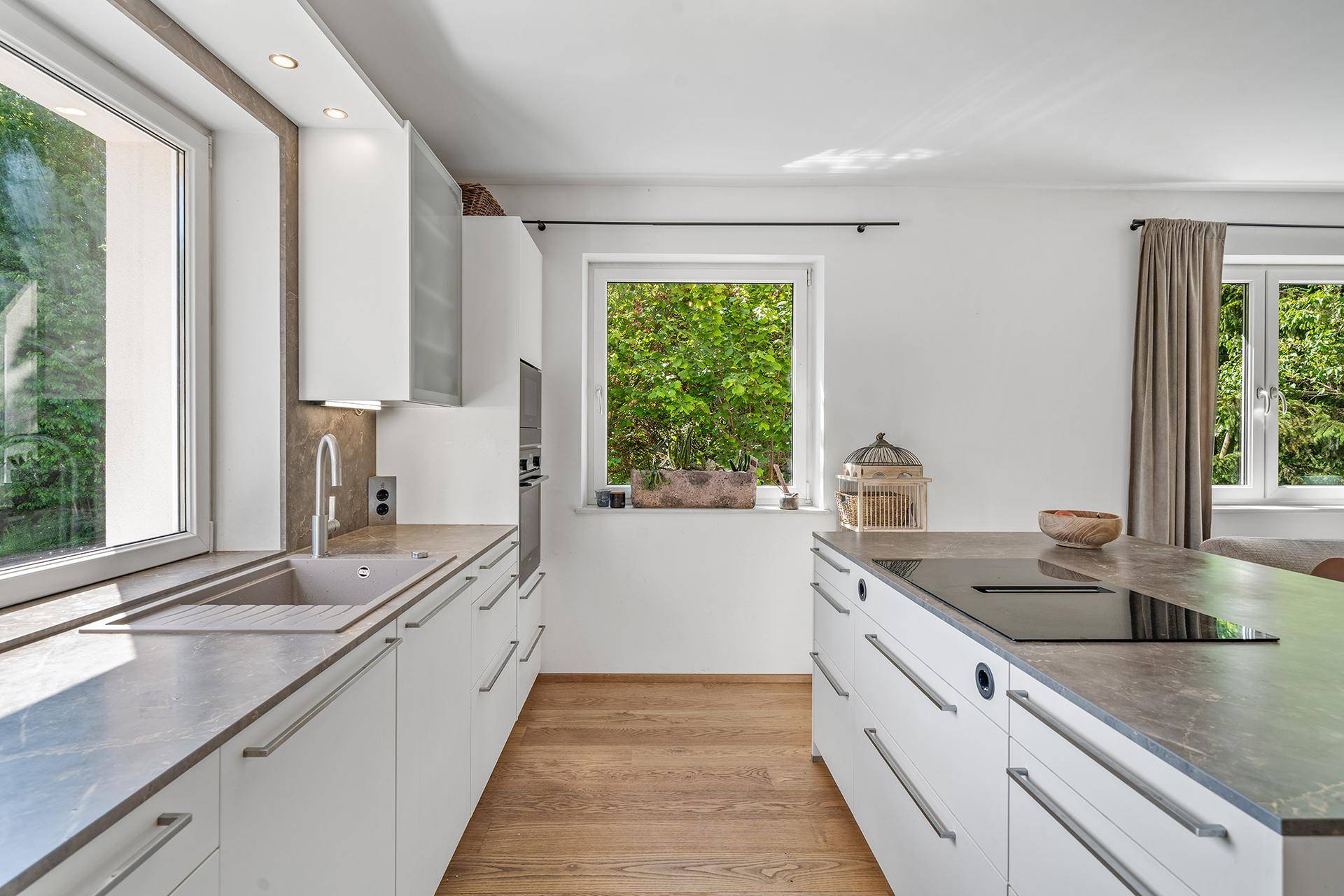 Moderne Küche mit weißen Schränken, dunkler Arbeitsplatte und Fensterblick ins Grüne.
