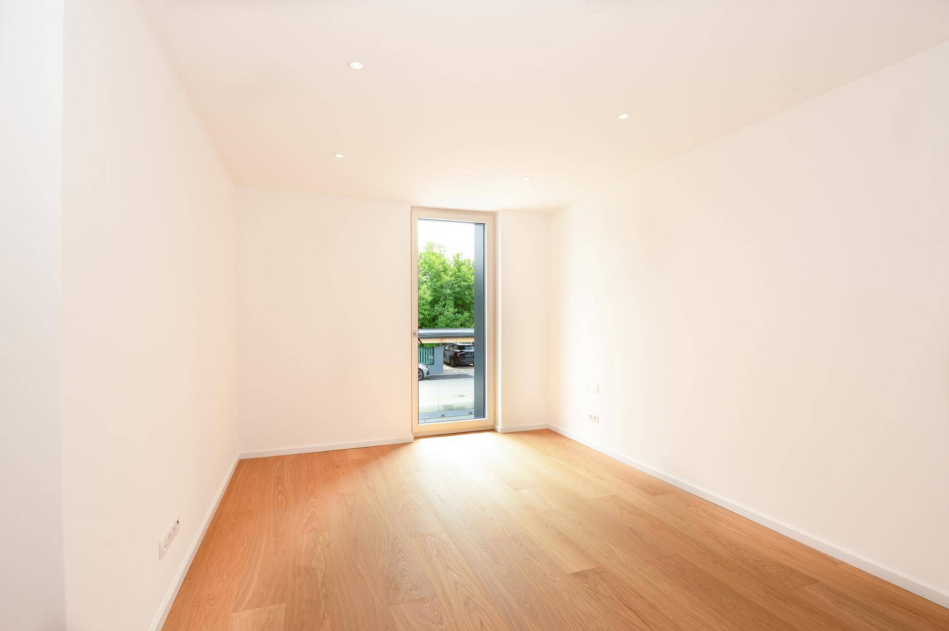 Helles Schlafzimmer mit Holzboden und einem hohen Fenster, das viel Licht hereinlässt.