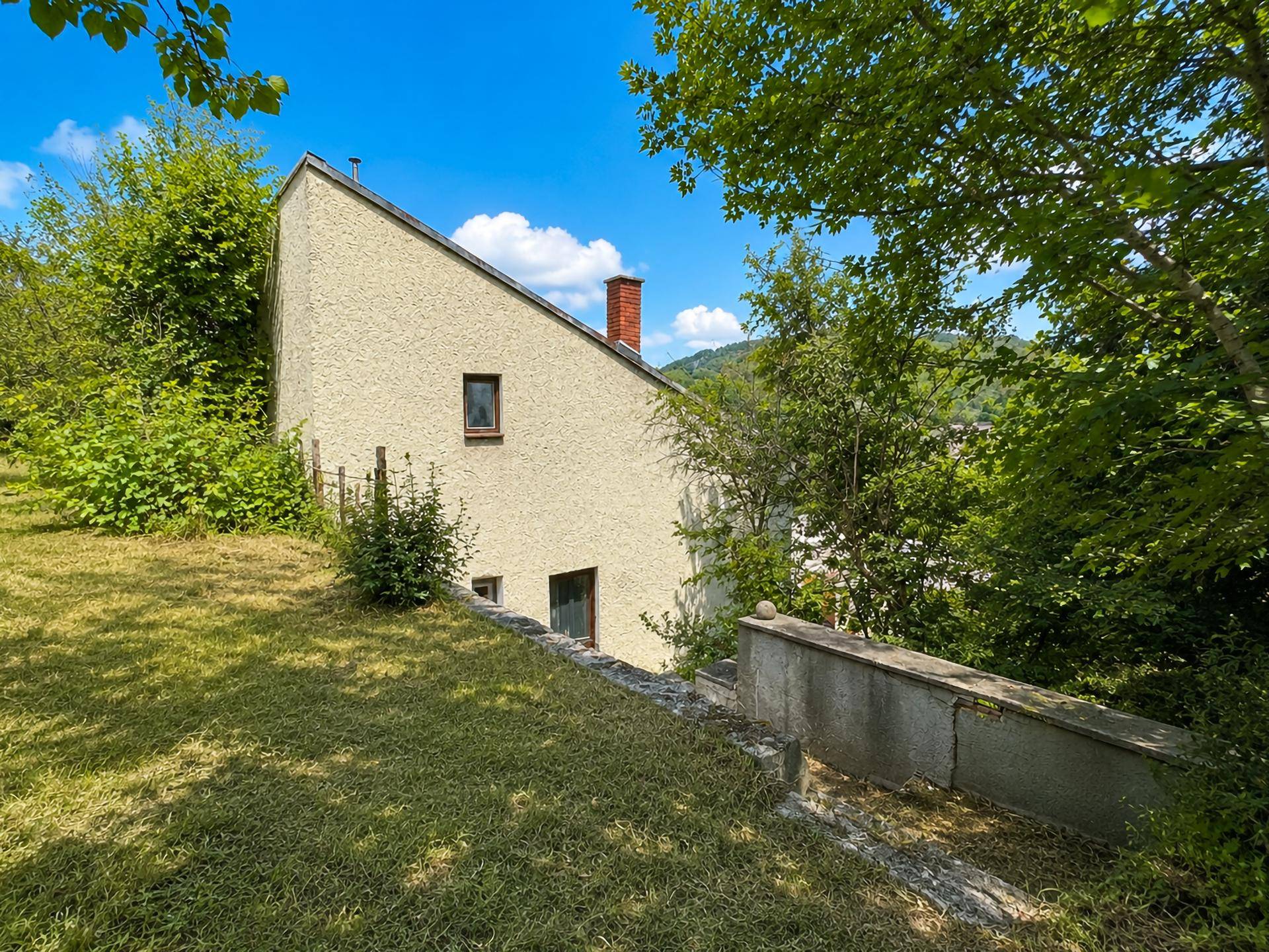 Seitenansicht eines Hauses mit schrägem Dach und Putzfassade, eingebettet in eine grüne Hanglage.