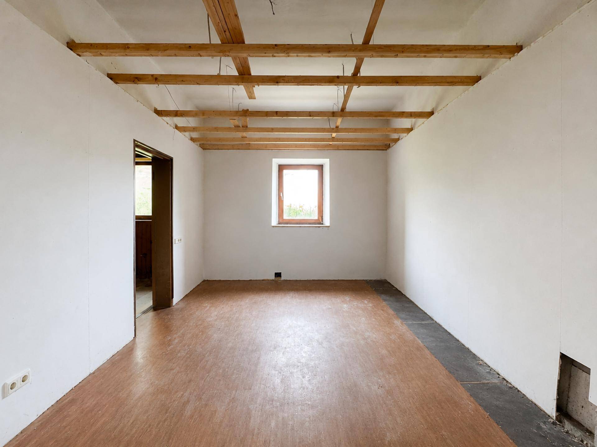 Leerer Raum mit freiliegenden Deckenbalken, weißen Wänden und einem Fenster, bereit zur Renovierung.