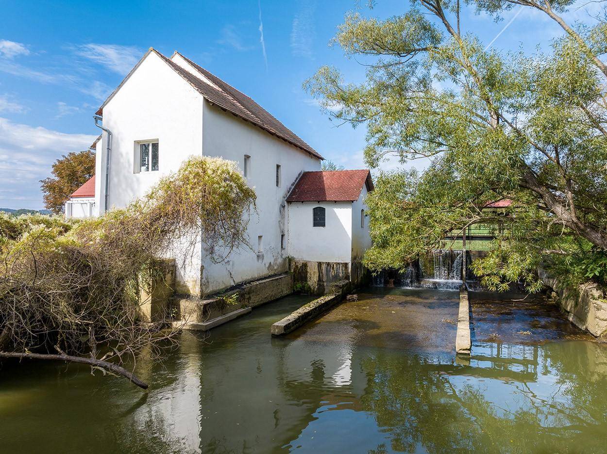Historisches Mühlengebäude am Wasserlauf mit kleinem Wasserfall und üppiger Vegetation.