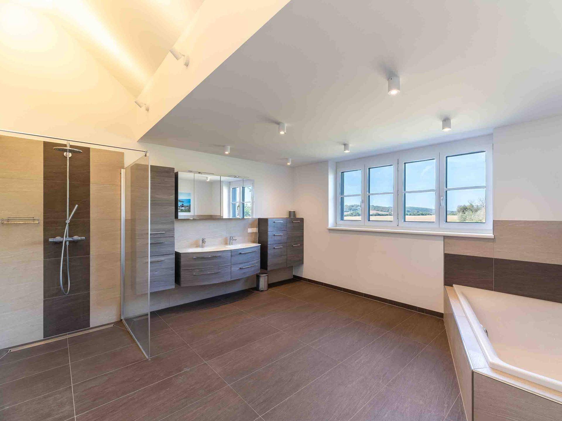 Geräumiges modernes Badezimmer mit Fliesenboden, großer Dusche, Doppelwaschbecken und Badewanne am Fenster.