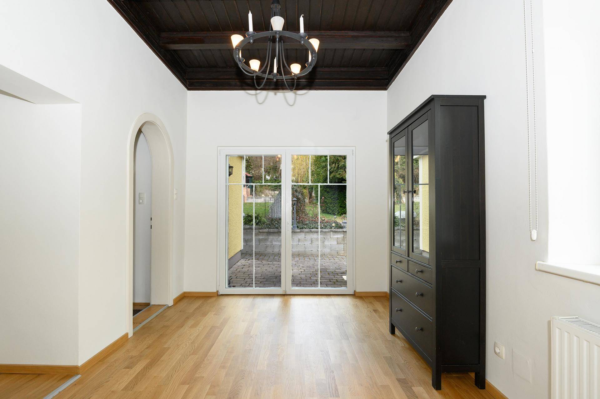 Geräumiger Raum mit dunkler Holzdecke und großen Fenstertüren zum gepflasterten Außenbereich.