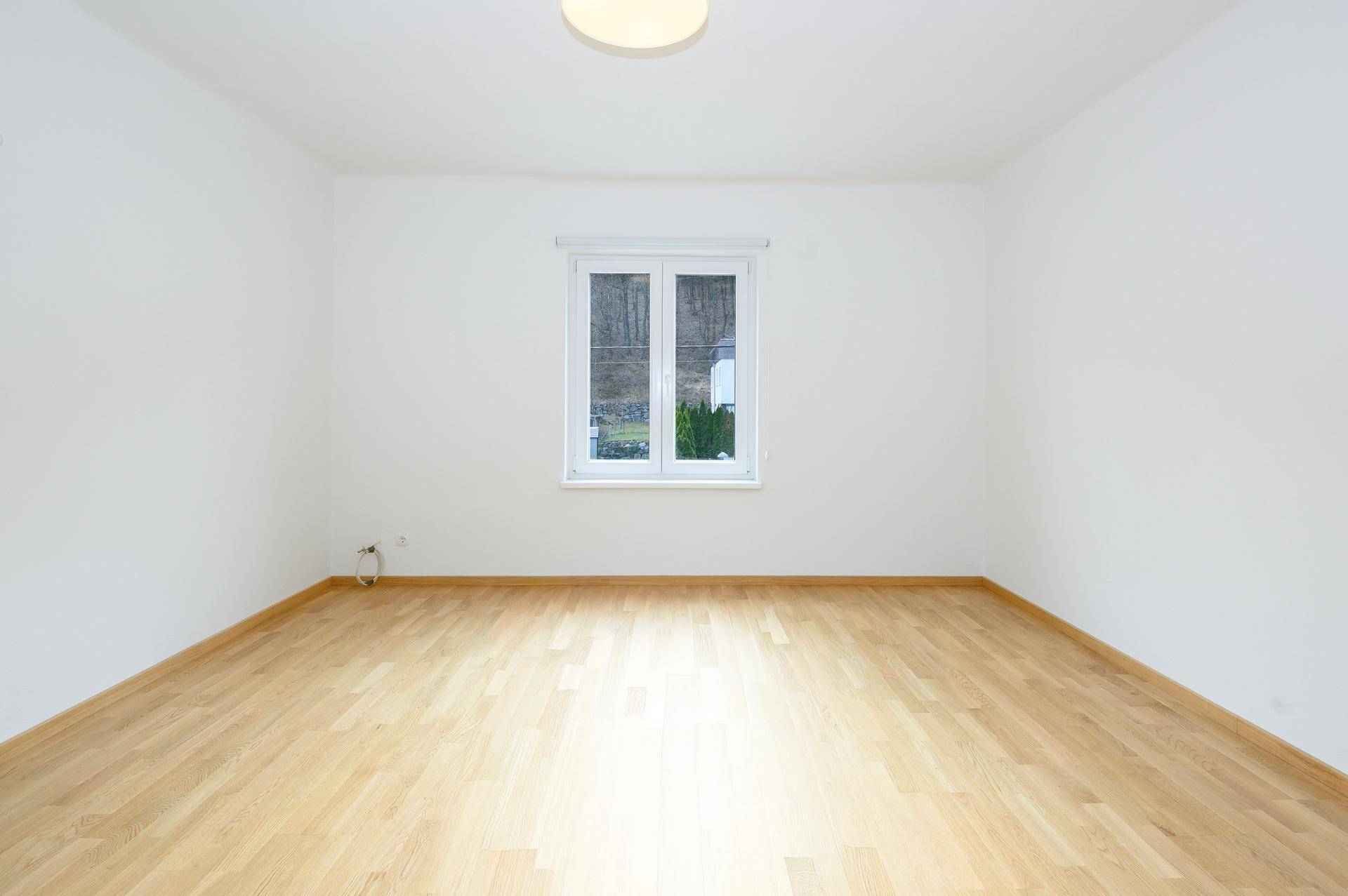Leeres Zimmer mit hellem Holzboden und einem Fenster, das viel Tageslicht hereinlässt.