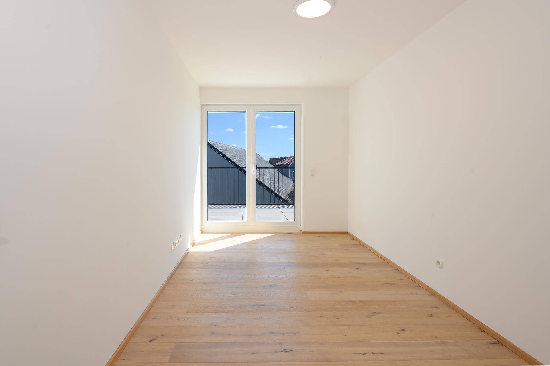 Lichtdurchflutetes Zimmer mit Holzboden und großen Fenstern, die Zugang zu einem Außenbereich bieten.