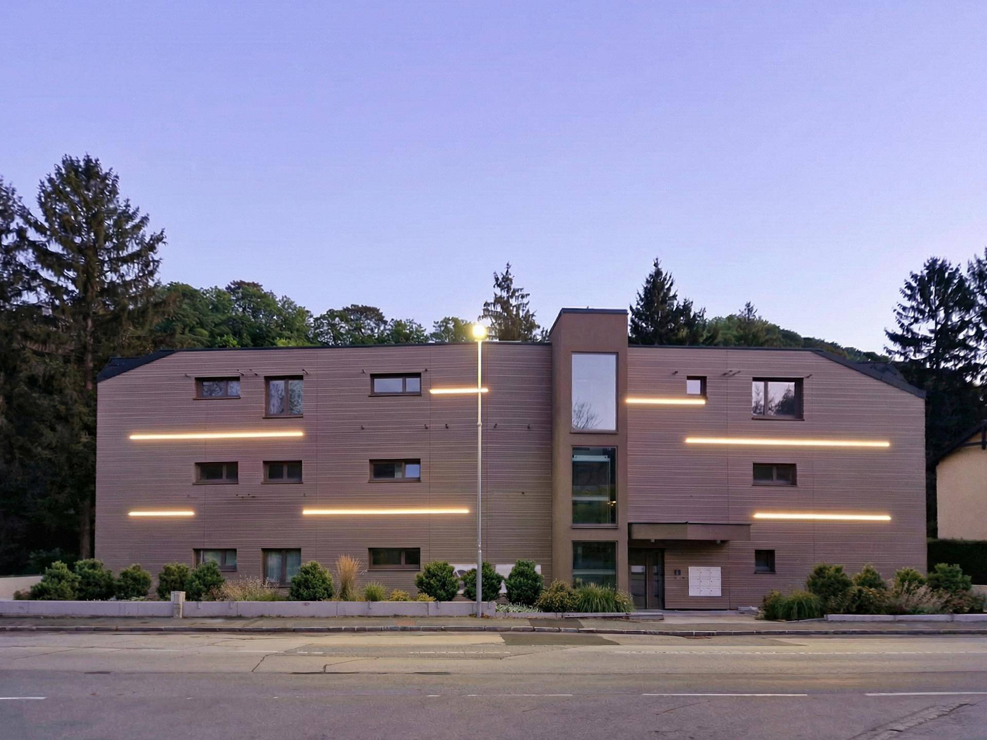 Moderne Fassade eines Wohngebäudes mit Holzverkleidung und integrierter Beleuchtung bei Dämmerung.