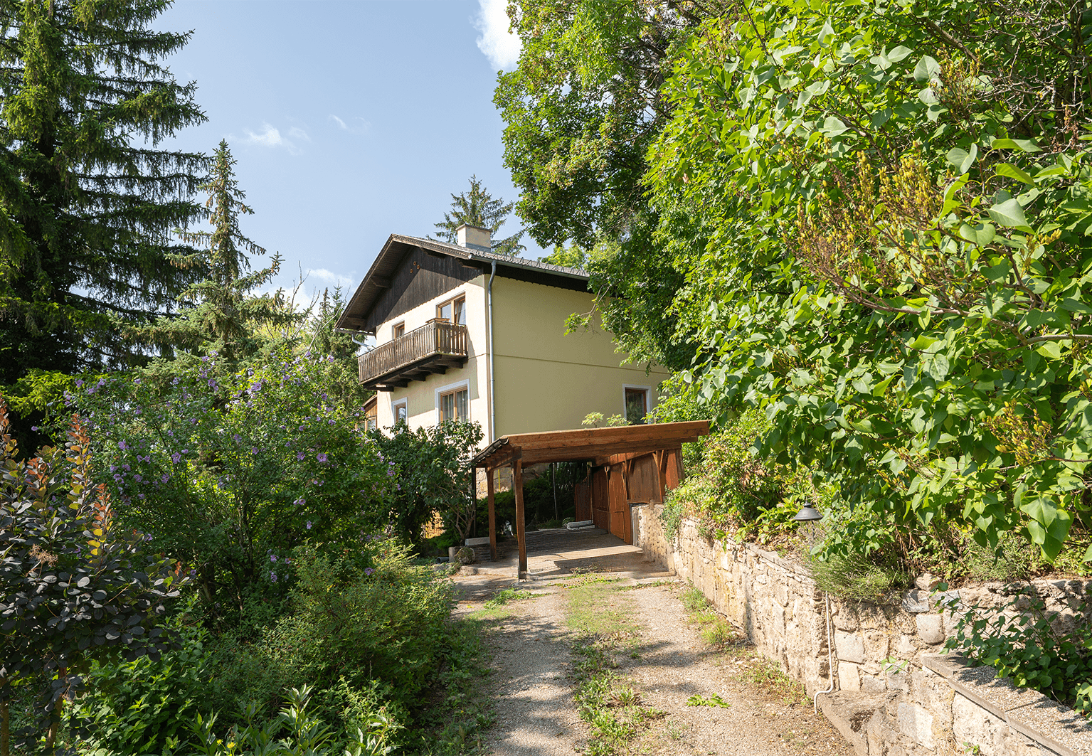 Ansicht des Hauses mit Carport und Zufahrt, umgeben von üppigem Grün.