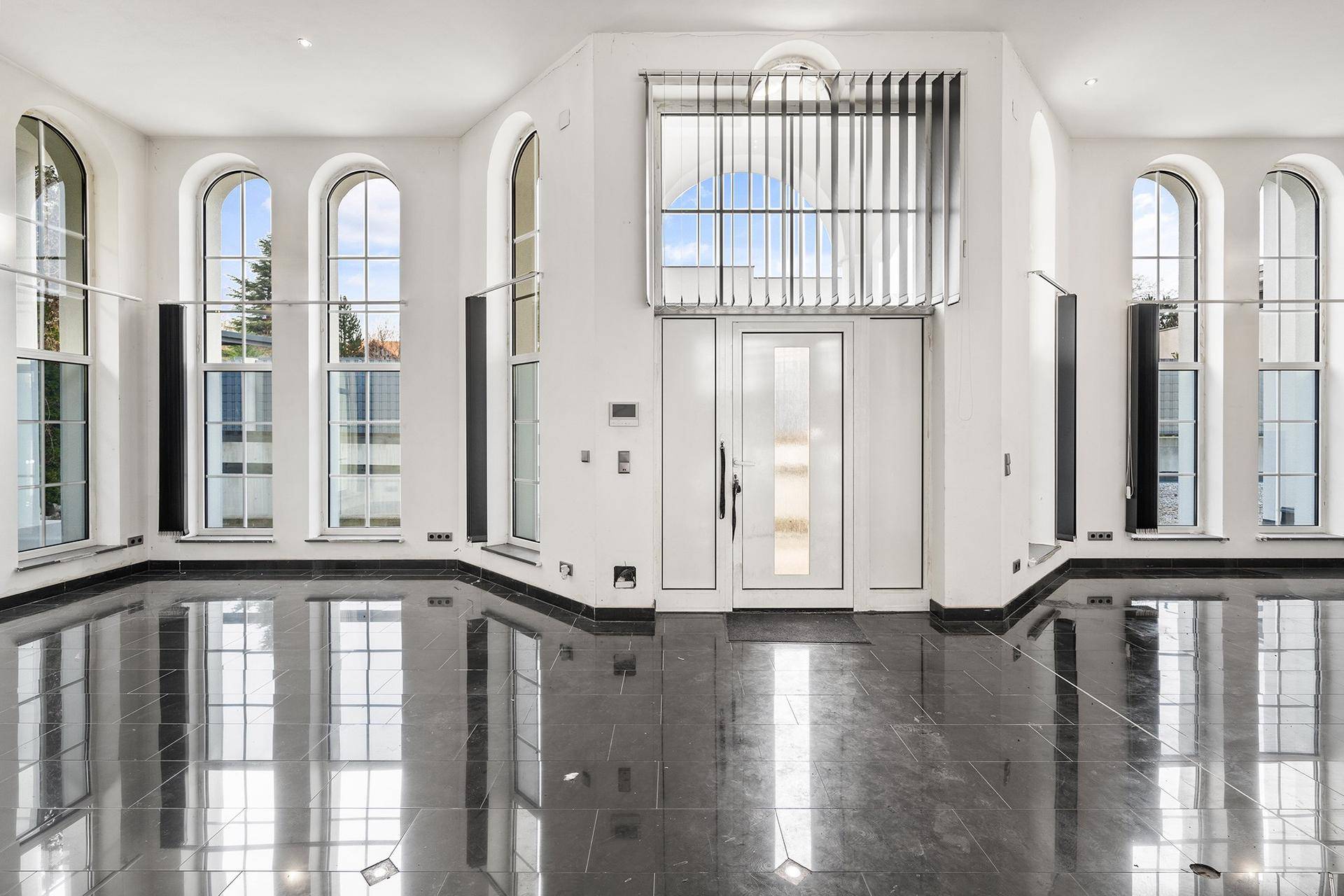 Heller Eingangsbereich mit hohen Bogenfenstern, glänzendem Fliesenboden und moderner Eingangstür.