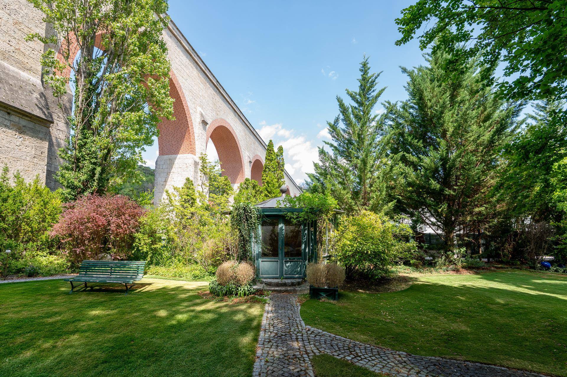 Gepflegter Garten mit Rasenfläche, Bäumen und einem Pavillon unter einem historischen Viadukt.