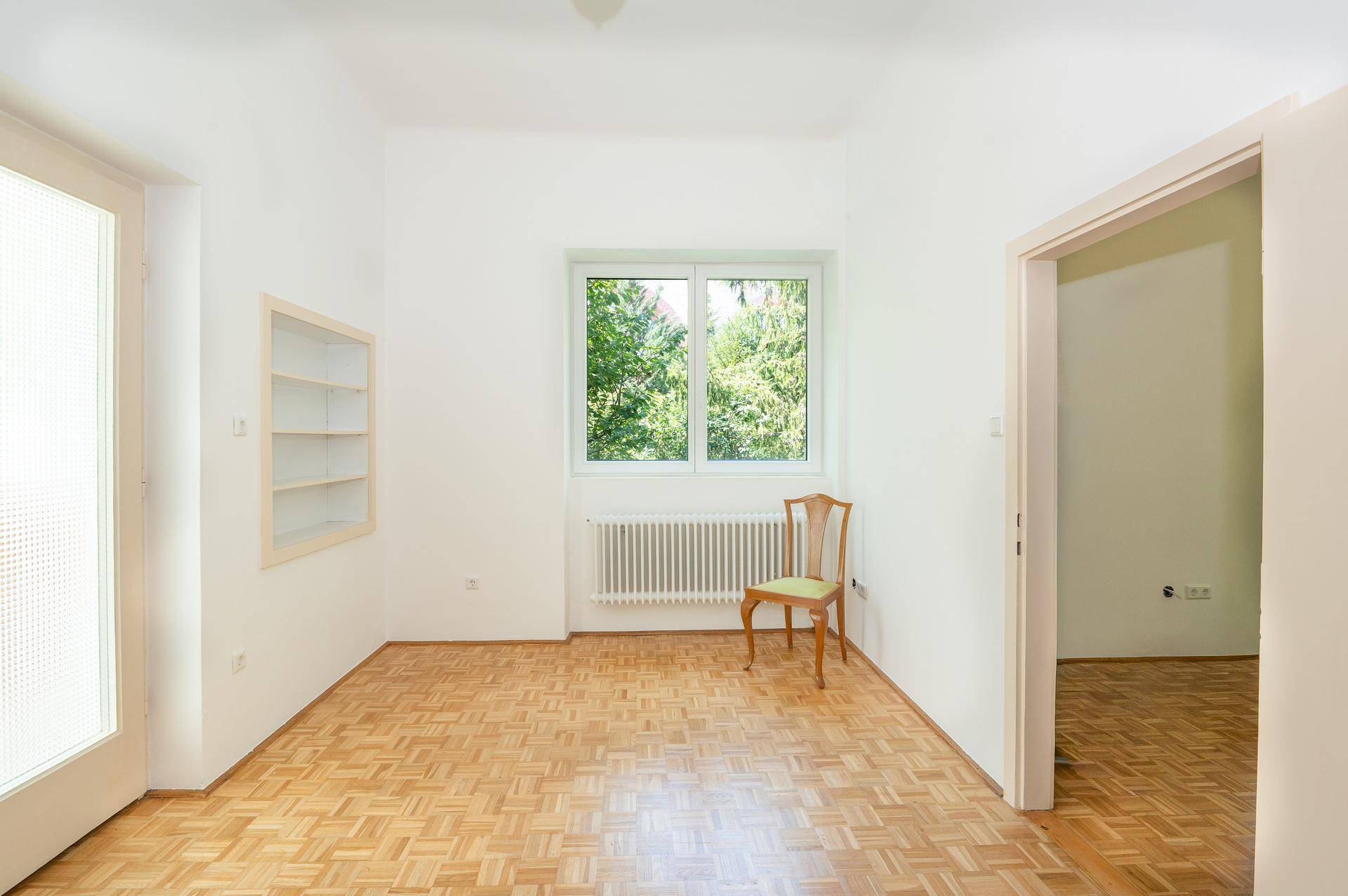 Leeres Zimmer mit Parkettboden, Fenster und eingebautem Regal, ideal als Büro oder Schlafzimmer.