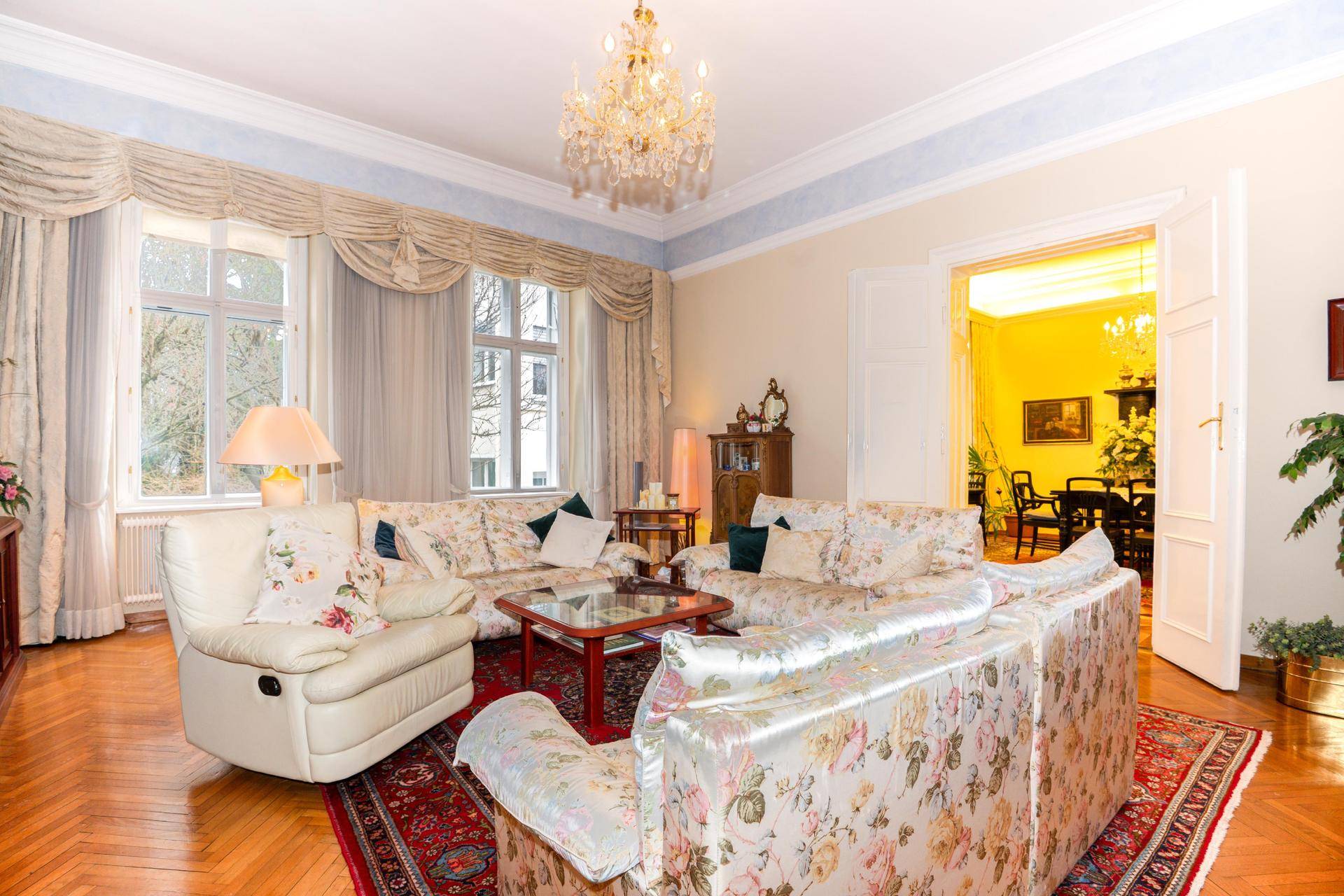 Großzügiges Wohnzimmer mit Parkettboden, floralen Sofas, Kronleuchter und hohen Decken.