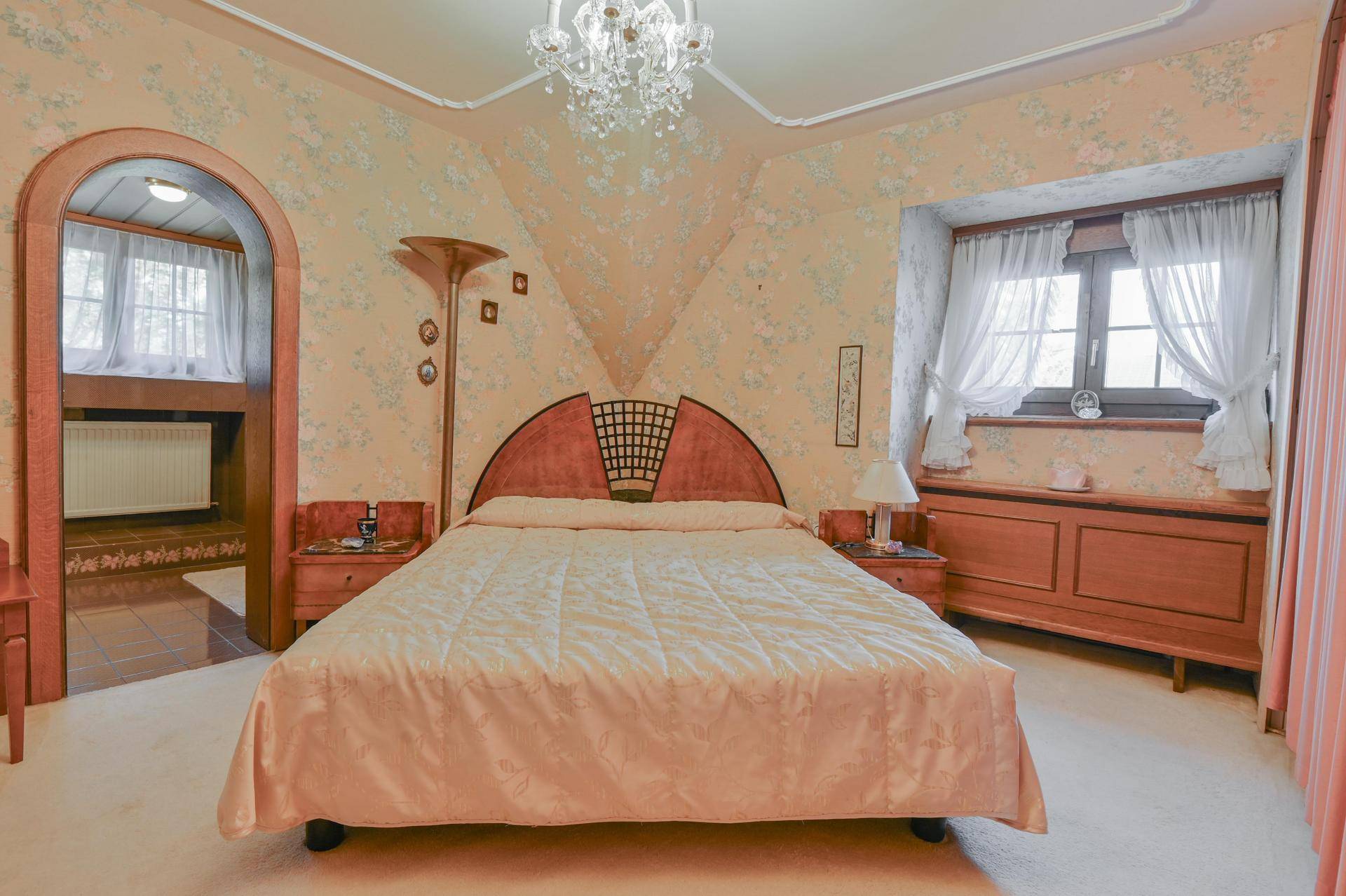 Traditionelles Schlafzimmer mit floraler Tapete, eleganten Möbeln und einem Bogenzugang zu einem Nebenraum.