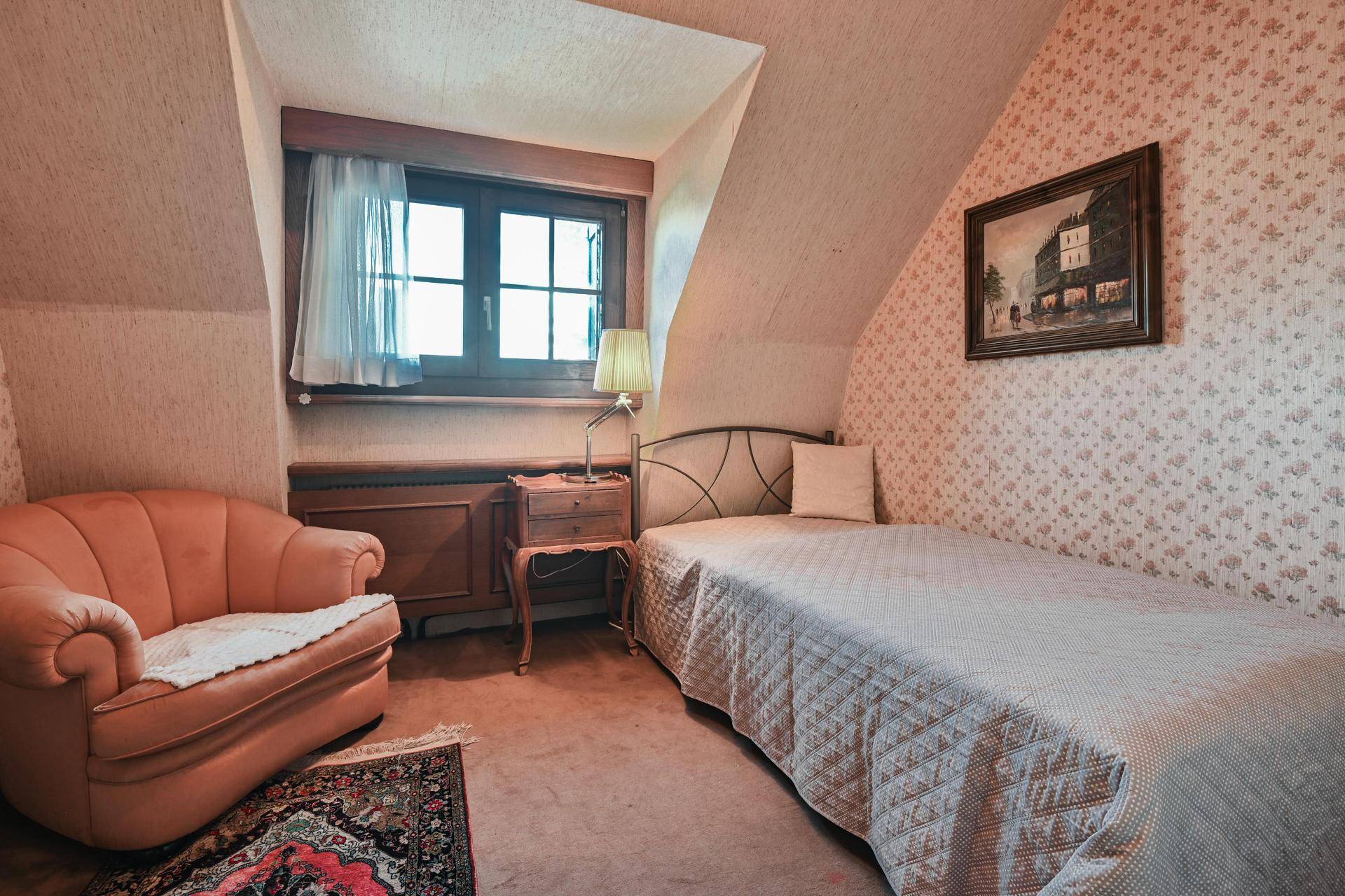 Gemütliches kleines Schlafzimmer mit Einzelbett, Sessel und floraler Tapete unter der Dachschräge.