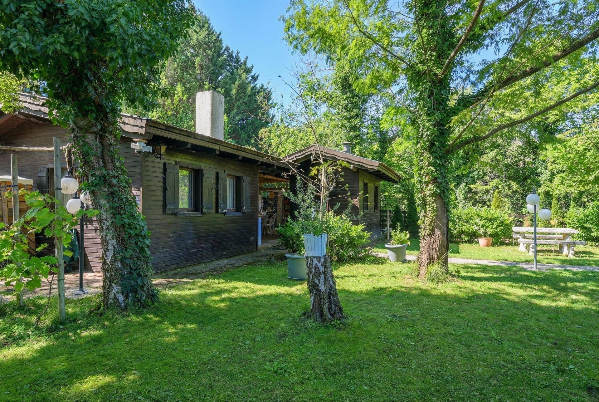 Großer Gartenbereich mit Holzhaus, alten Bäumen und einer Steinbank, umgeben von Natur.
