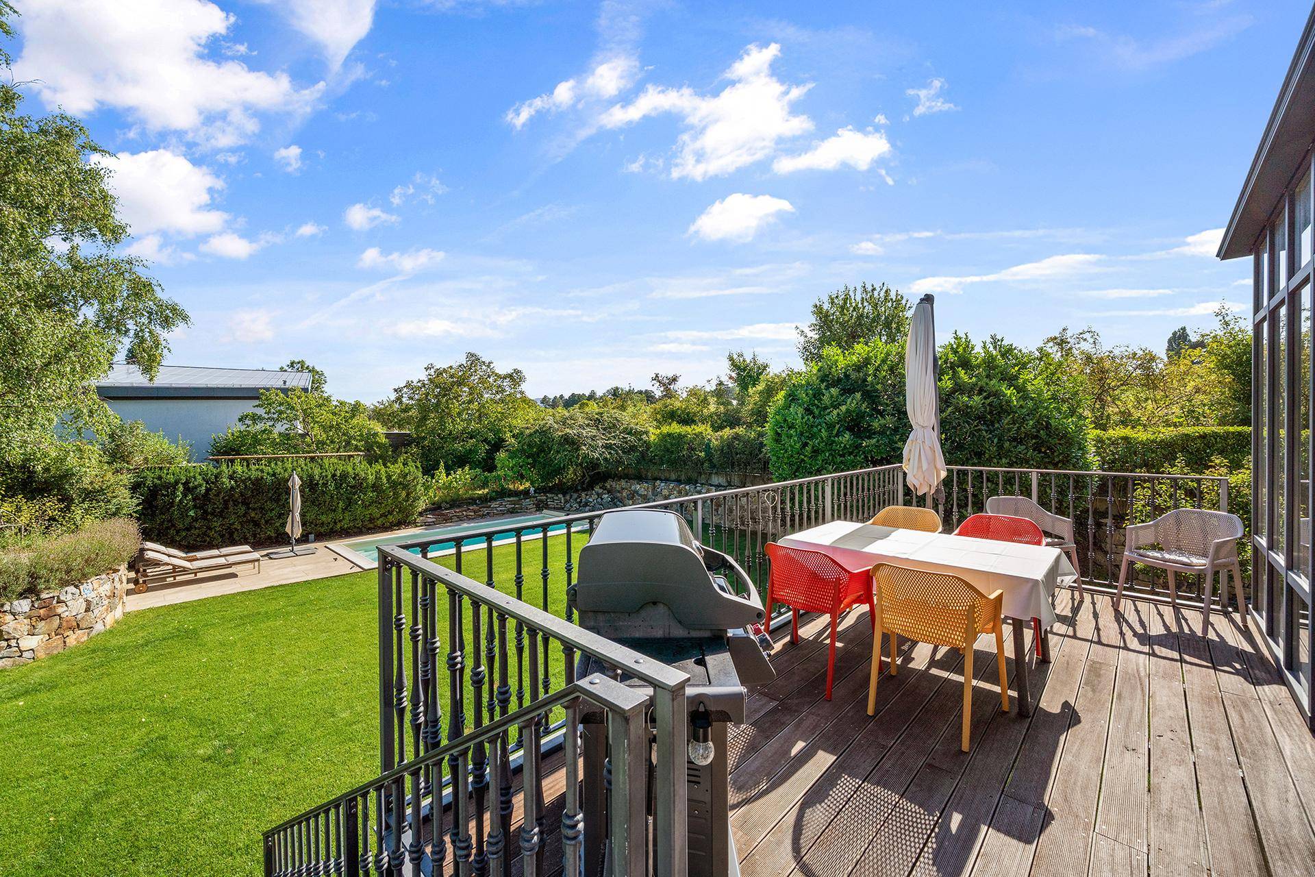 Holzterrasse mit Grillbereich und Esstisch im Freien, mit Blick auf Garten und Pool.