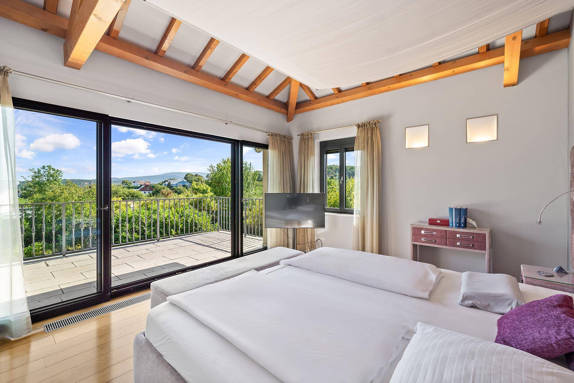 Helles Schlafzimmer mit Holzbalken, Balkonzugang und weitem Blick über die Landschaft.