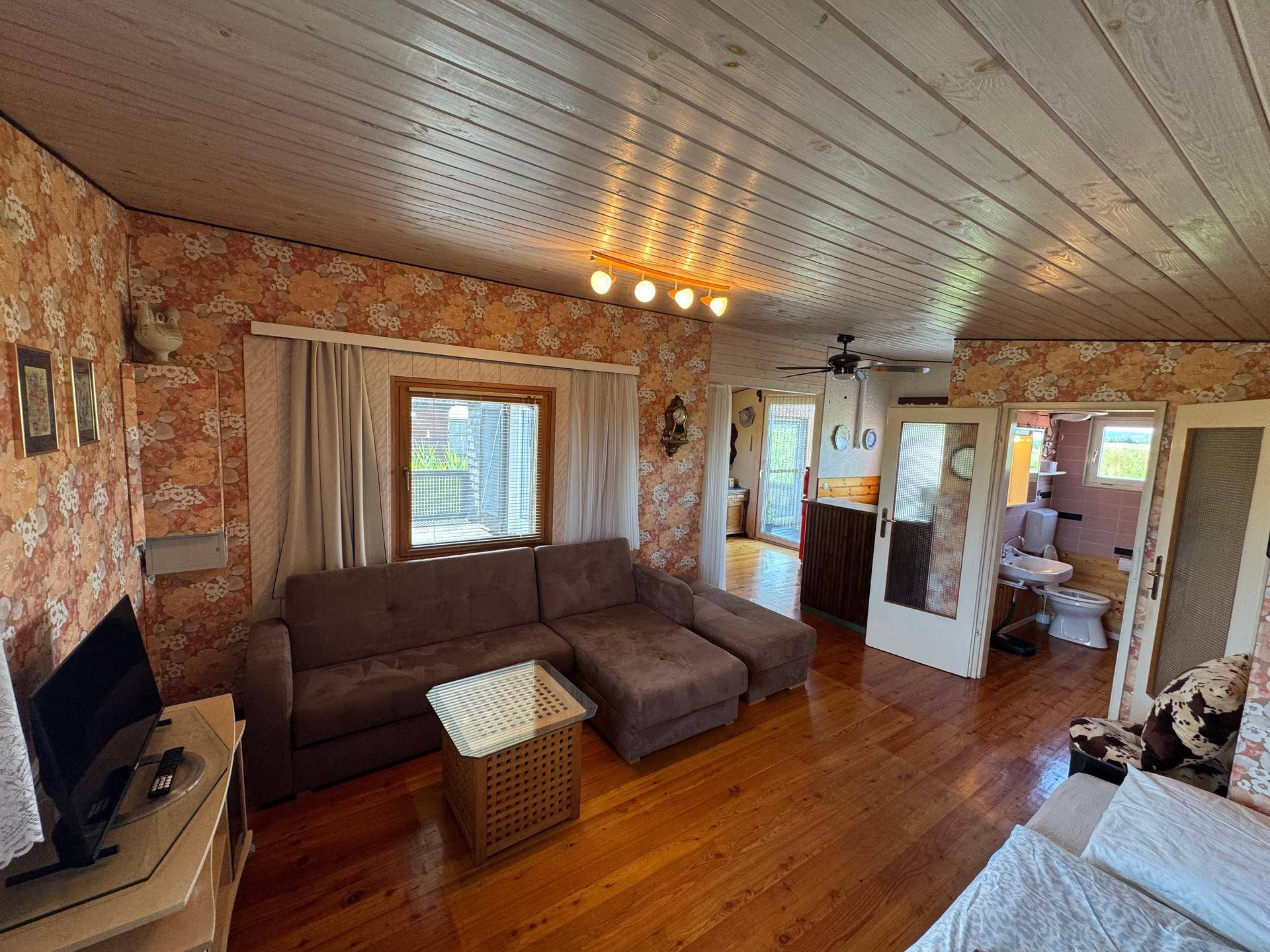 Gemütliches Wohnzimmer mit großem Sofa, Holzboden und floraler Tapete, mit Zugang zum Bad.