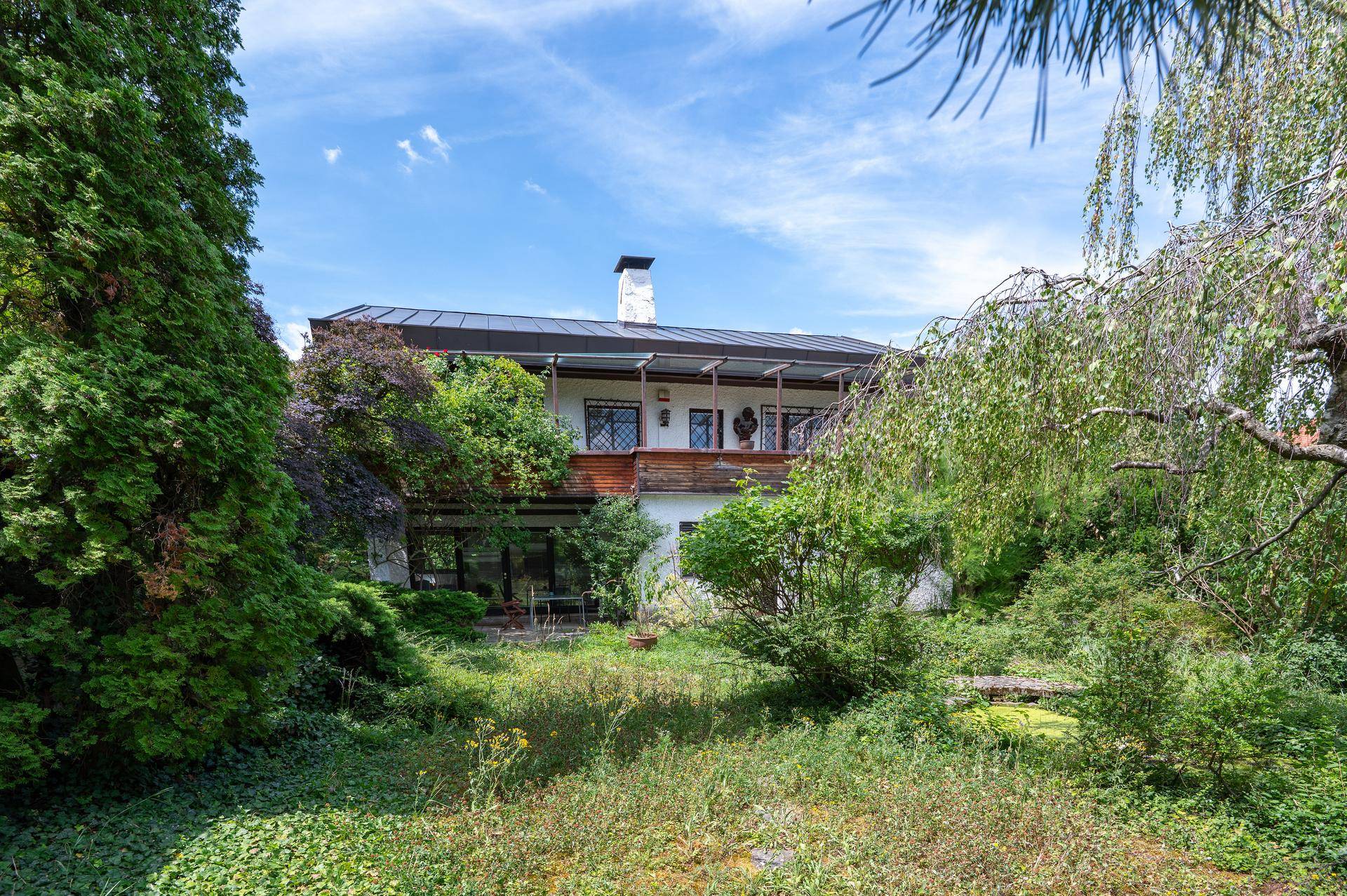 Ansicht des Hauses von außen, umgeben von üppigem Grün und hohen Bäumen unter blauem Himmel.